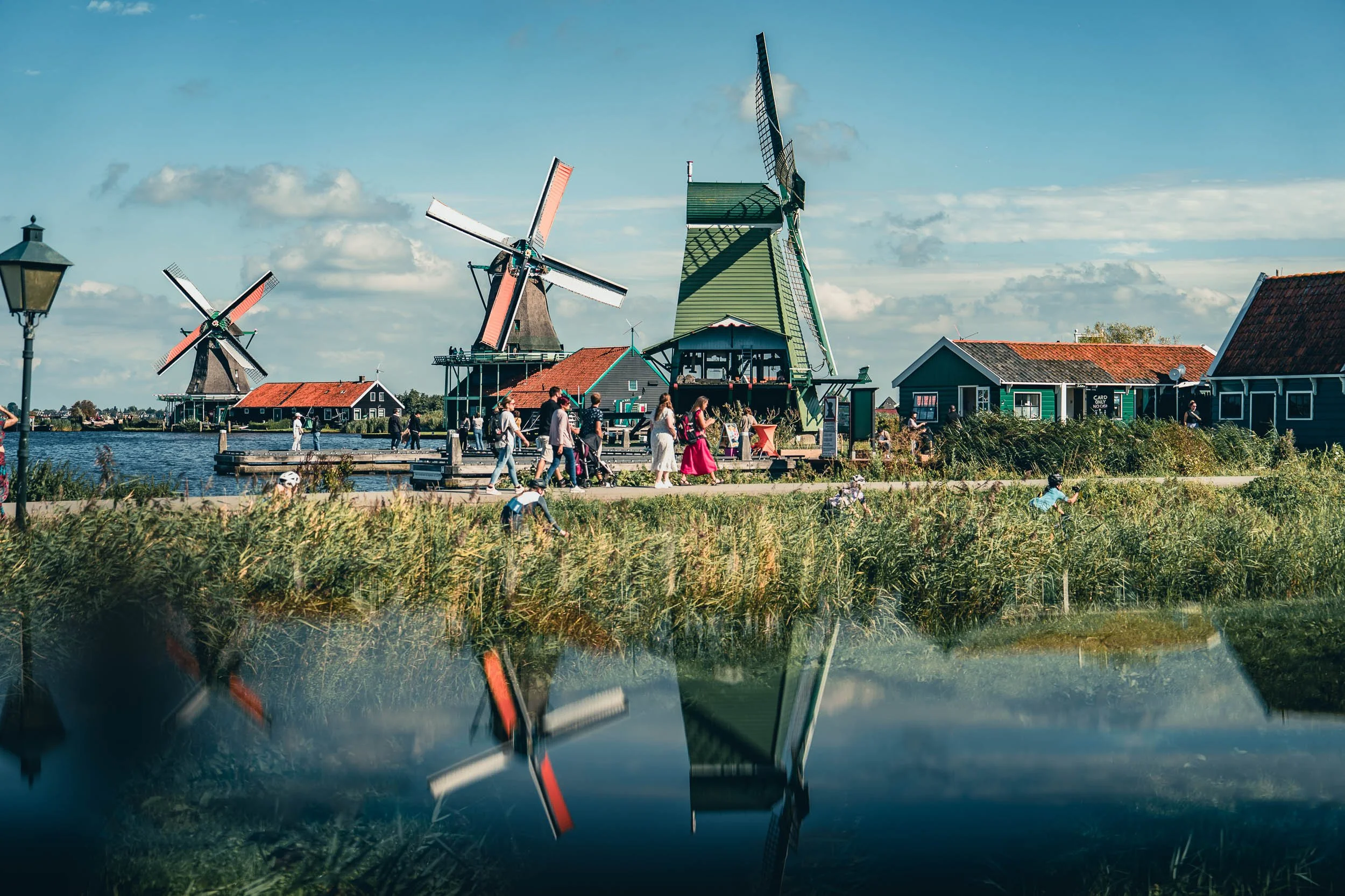 A scenic view of traditional windmills near a water body with people walking and biking along the pathway, under a partly cloudy sky.