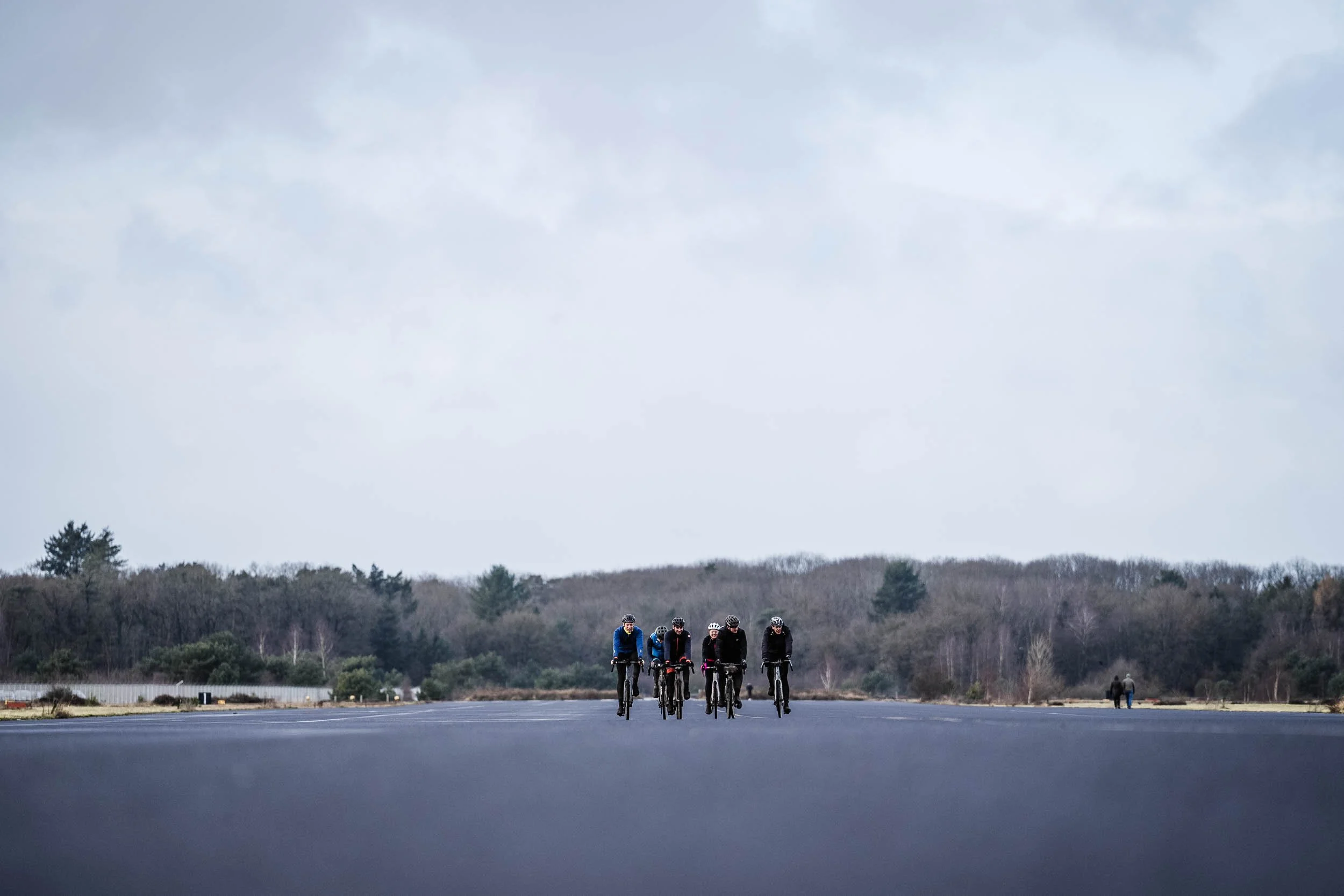 Five cyclists riding on an empty road with a forested landscape in the background, overcast sky.