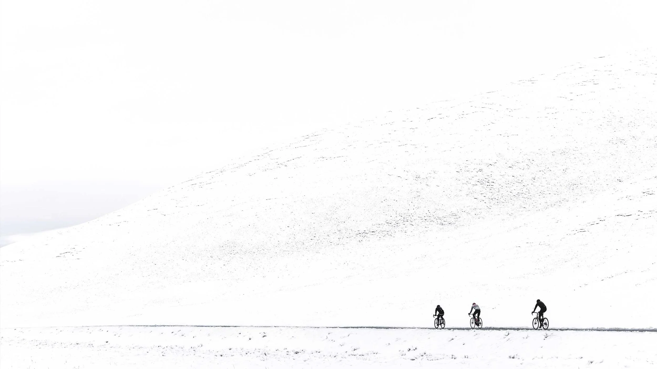 Three people riding bicycles on a snow-covered landscape with a large snowy hill in the background.