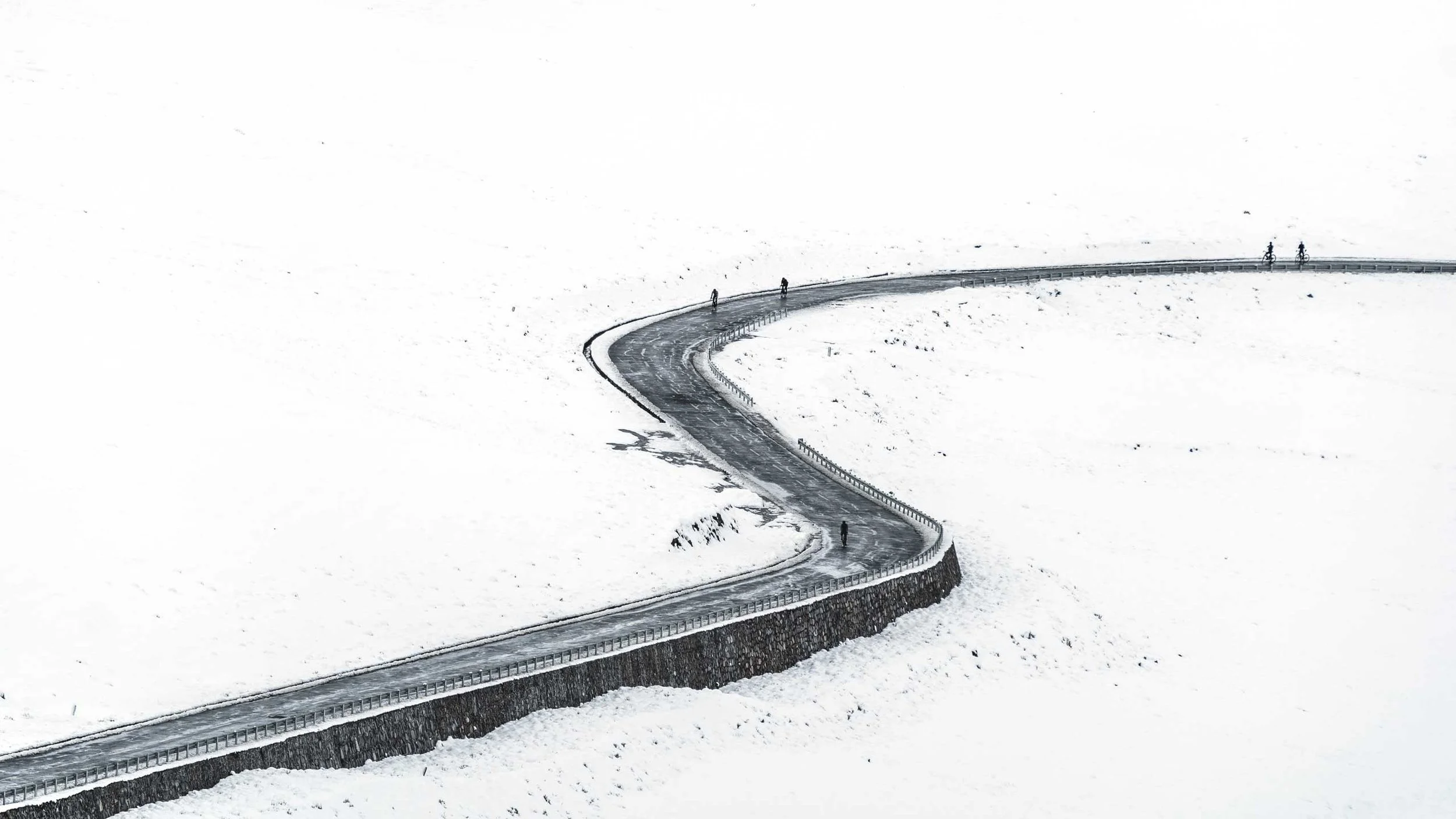 A winding road in a snowy landscape with snow-covered ground and four people walking or riding bikes along the road.