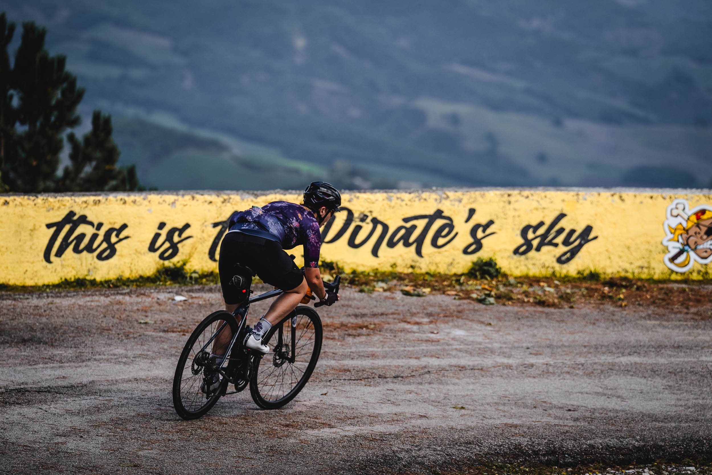 Cyclist riding on a mountain road with a yellow wall behind them that has black text and cartoon character artwork