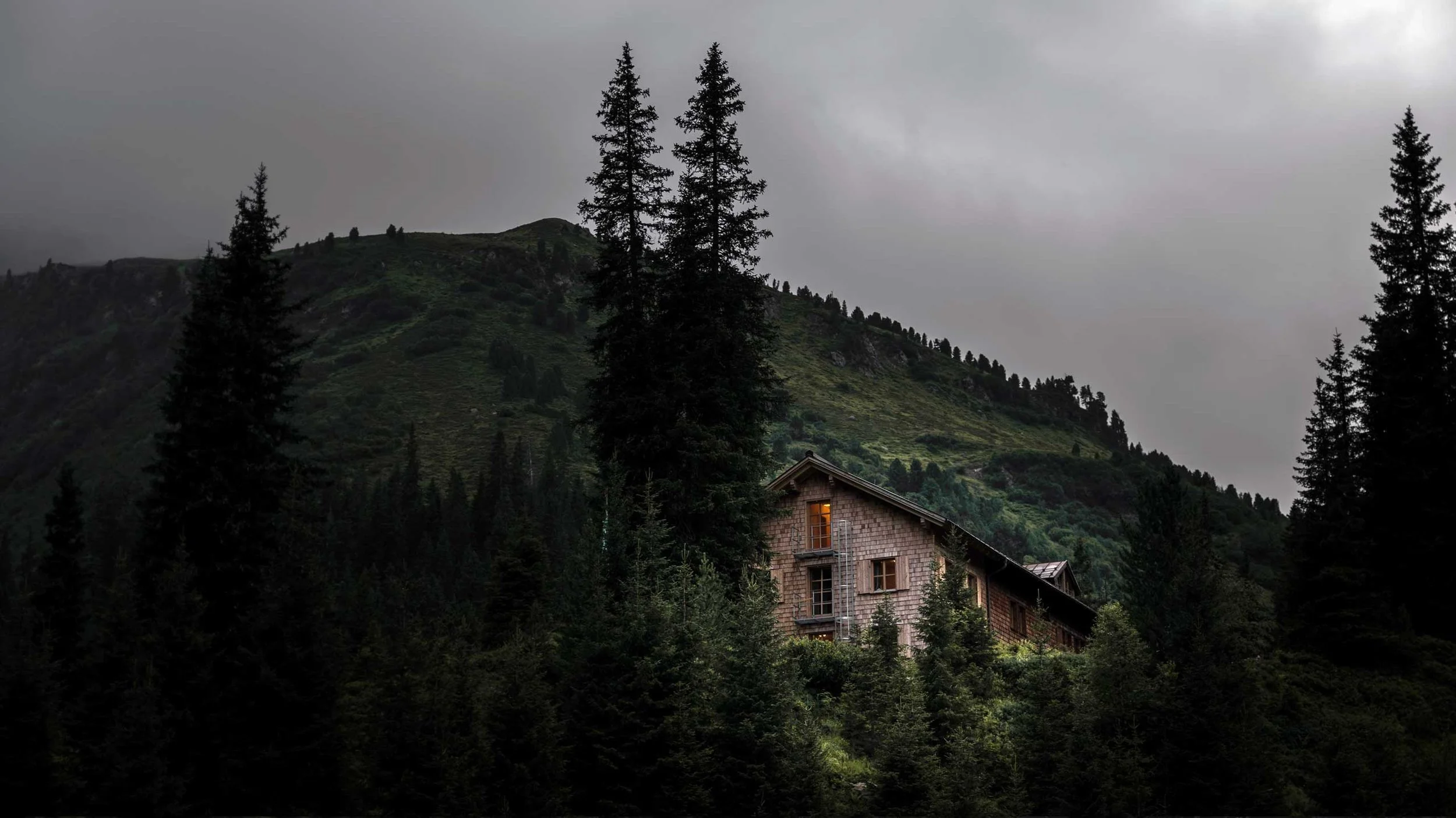 A house surrounded by tall pine trees on a hillside with mountains in the background under a cloudy sky.