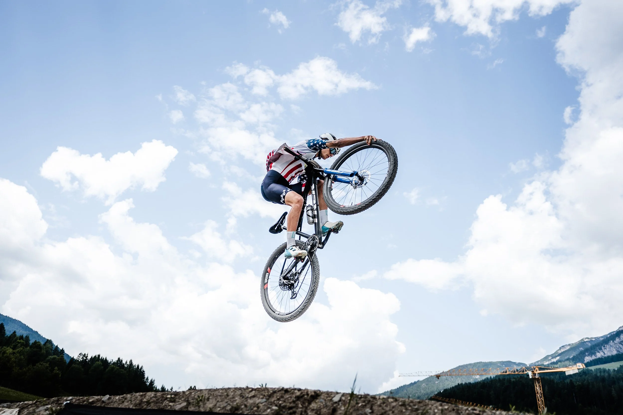 A person wearing patriotic clothing is mid-air on a mountain bike, performing a jump outdoors against a blue sky with scattered clouds and mountainous landscape in the background.