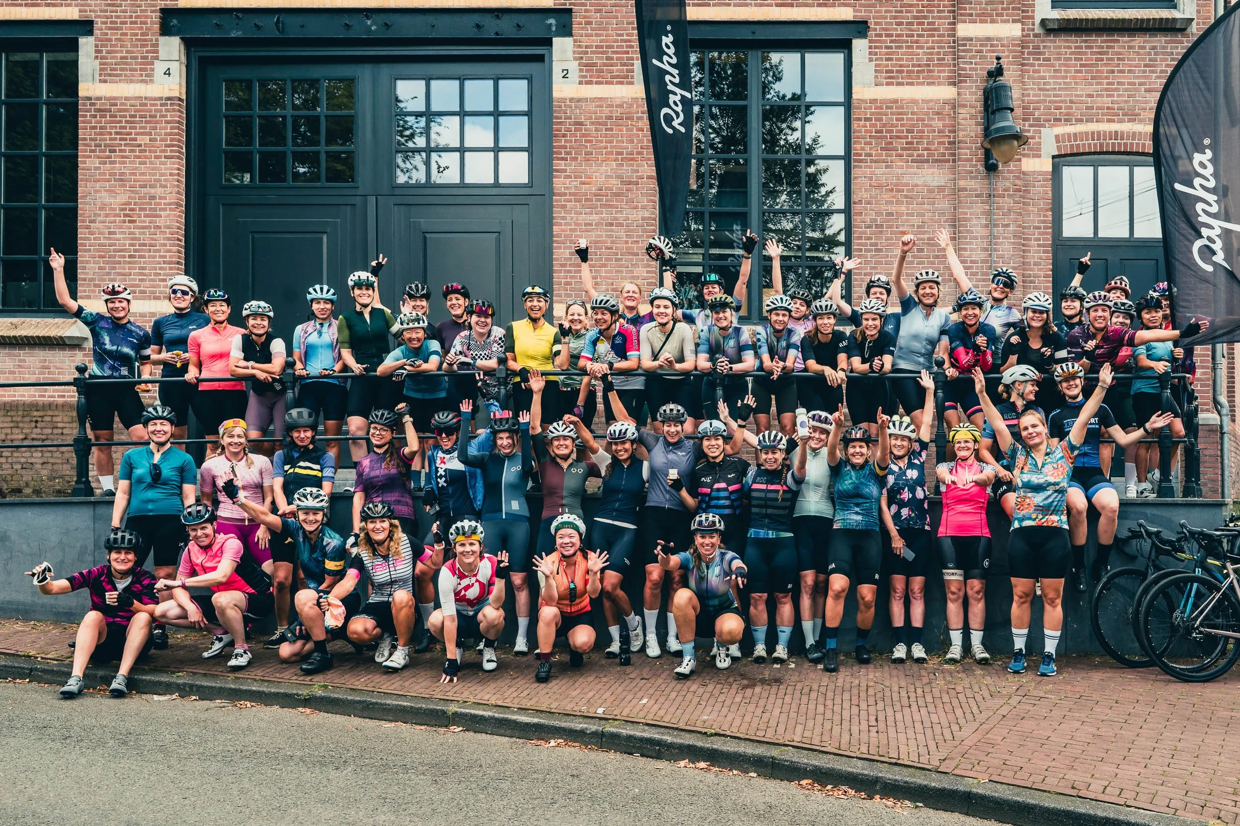 Group of women of various ages in cycling gear, helmets, and gloves posing together in front of a brick building with large windows during daytime. Some women are standing on steps, others are crouching or sitting, and they appear excited and cheerfu