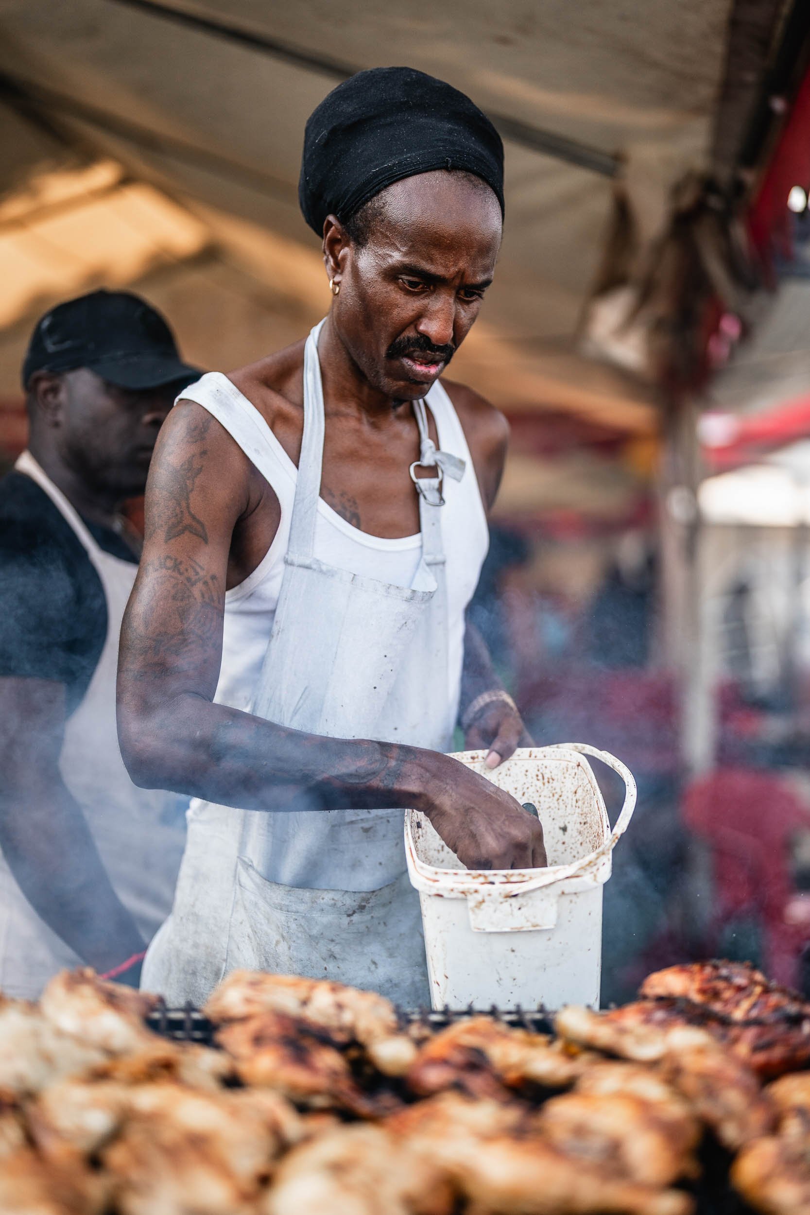 A man cooking meat at an outdoor grill, wearing a white apron and a black head covering, with another person in the background. (Patta.nl Rapha)