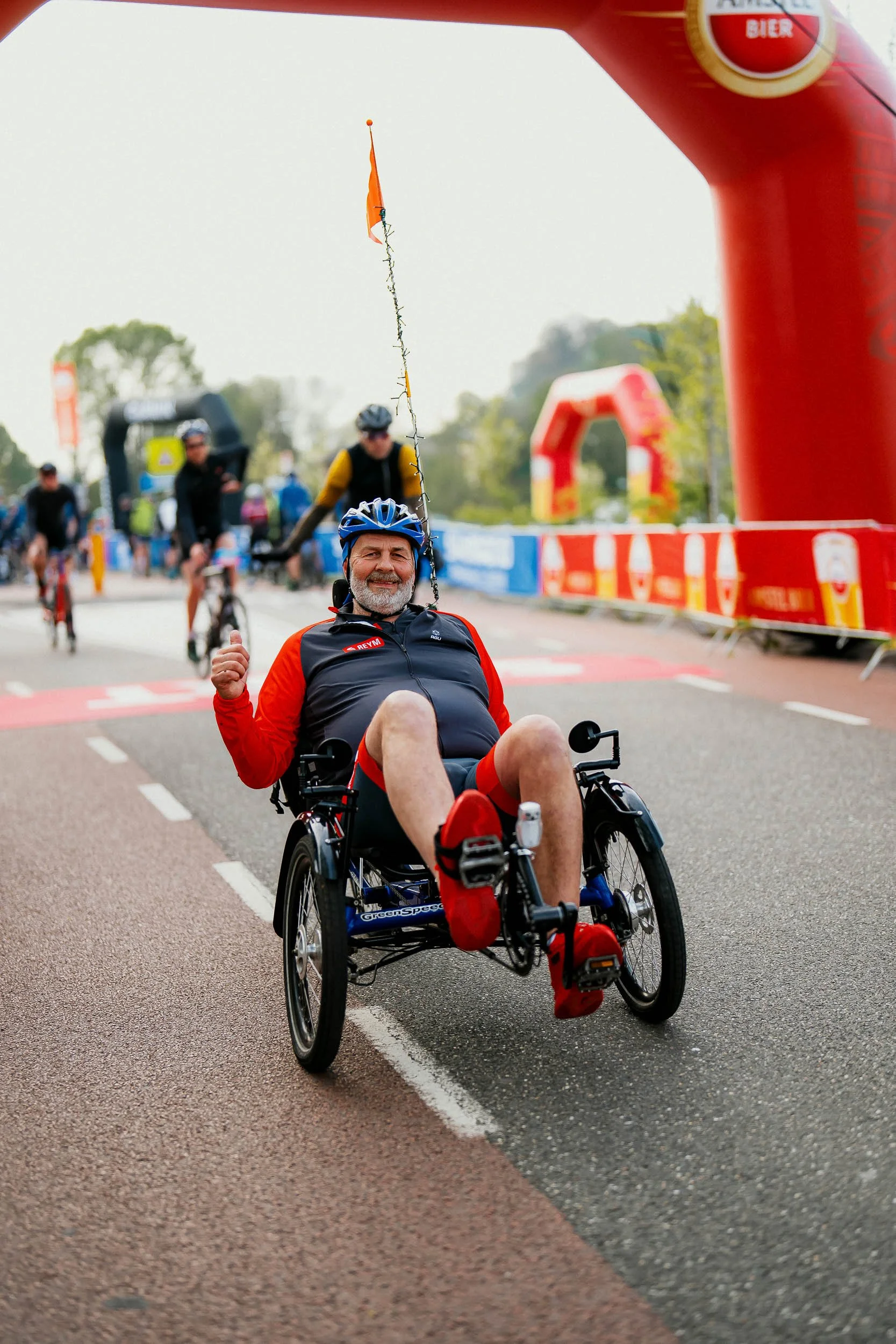 Smiling man with a beard in a recumbent tricycle crossing a race finish line, giving a thumbs-up gesture, wearing a blue helmet, black and red cycling gear, at an outdoor cycling event with other cyclists and race banners in the background.