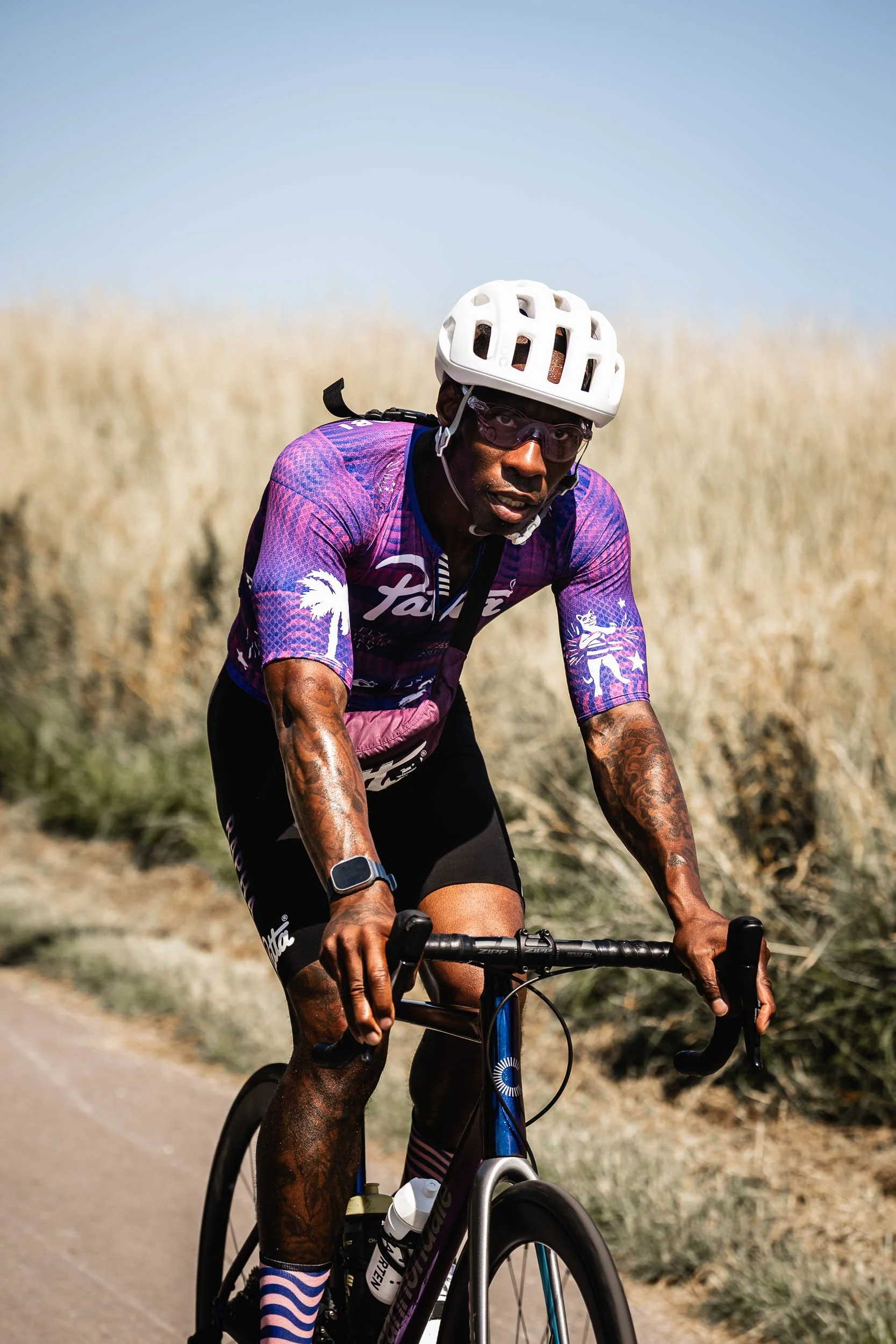 A male cyclist wearing a purple and pink cycling jersey, black shorts, a white helmet, and sunglasses, riding a bike on a path through a dry grass landscape on a sunny day.