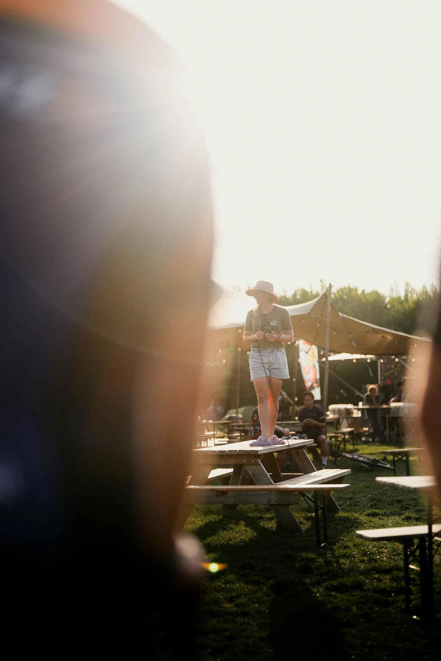 A woman wearing a wide-brimmed hat, shorts, and a t-shirt standing on a picnic table at an outdoor event, holding a microphone, with tents and other people in the background. (Graveller) )(Canyon)