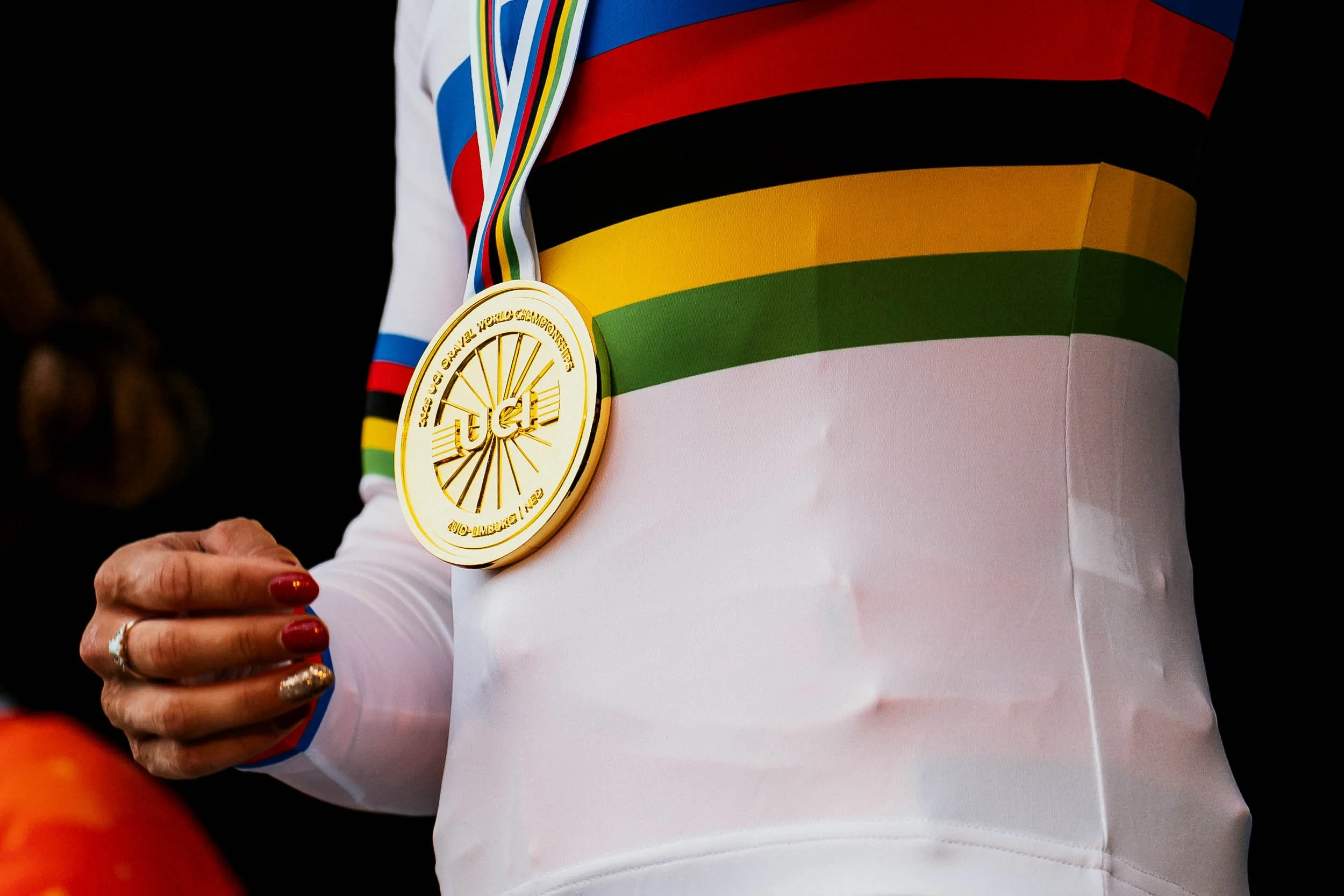 Close-up of a cyclist's white jersey with rainbow stripes and a gold medal hanging from a rainbow-colored ribbon around the neck, with a black background.