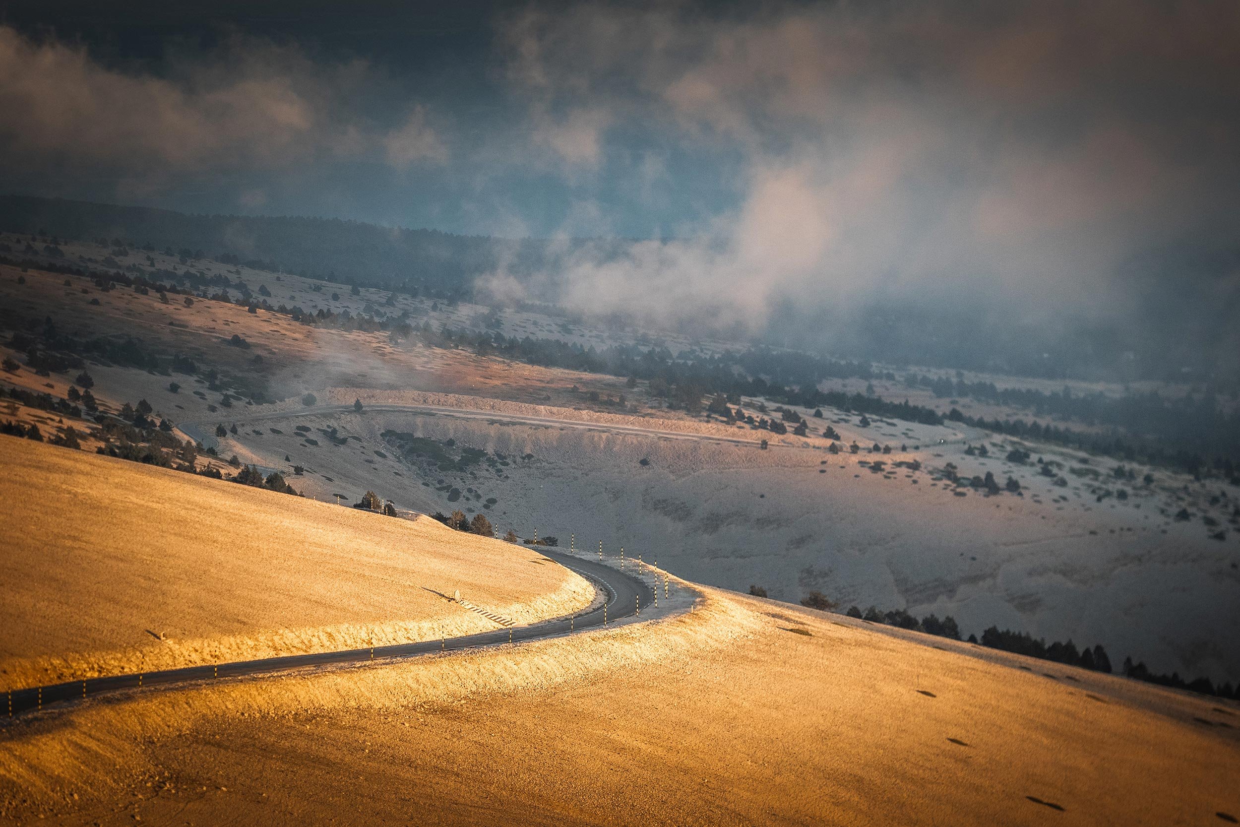 A winding road through rolling yellow and brown hills with a backdrop of mountains and partly cloudy sky.