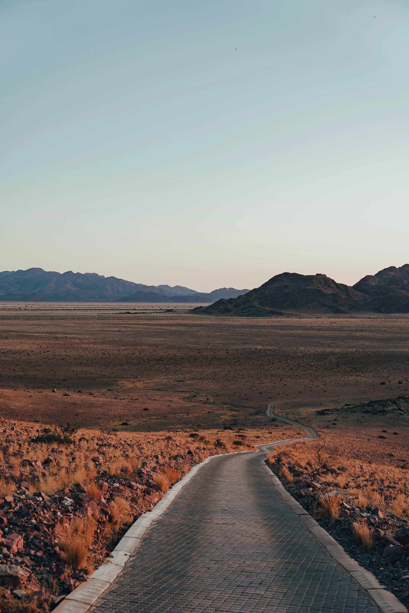 A winding paved road in a desert landscape with mountain ranges in the distance under a clear sky.
