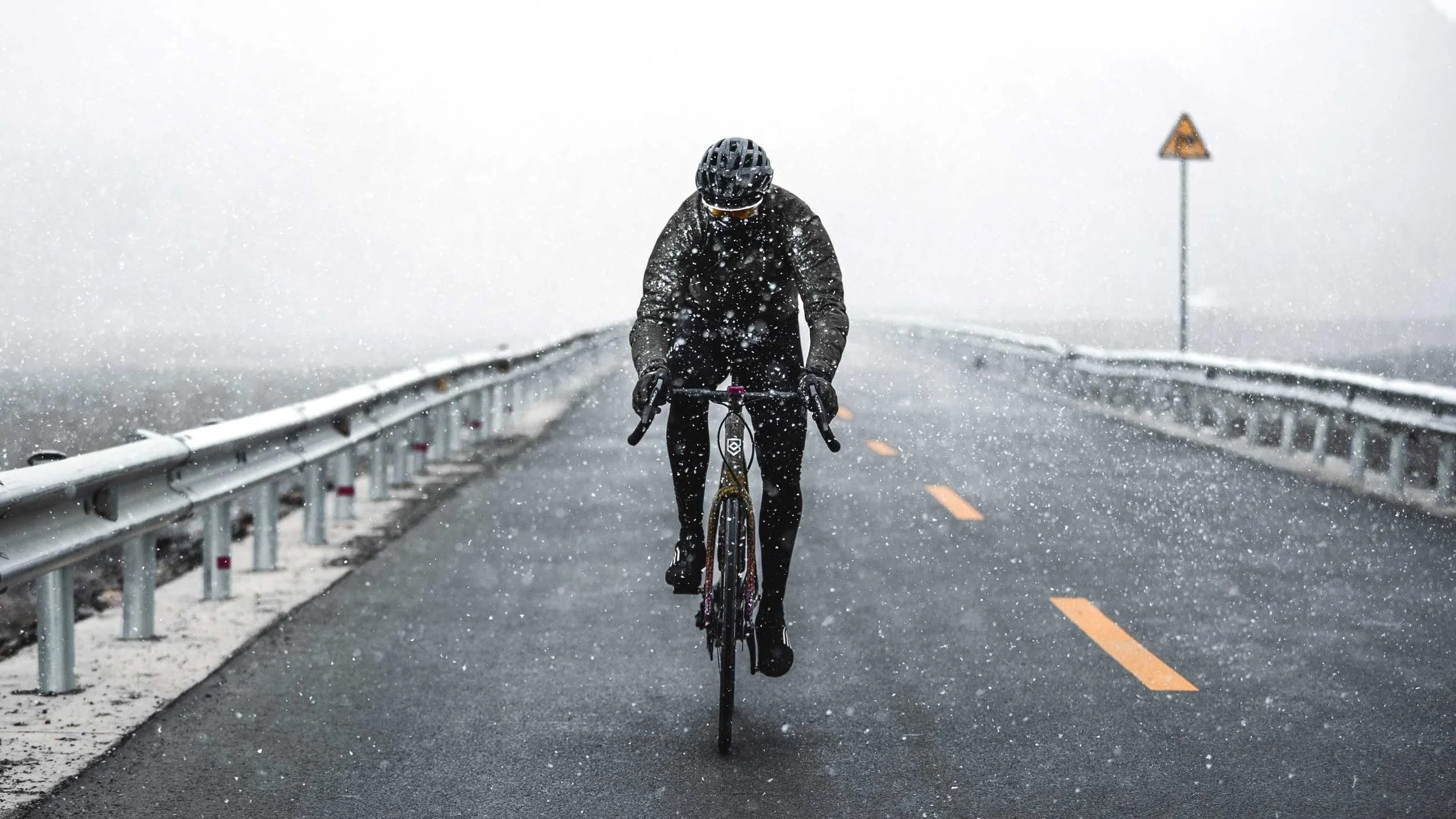 Person riding a bicycle in the rain or snow on a mountain road with guardrails and a foggy background.