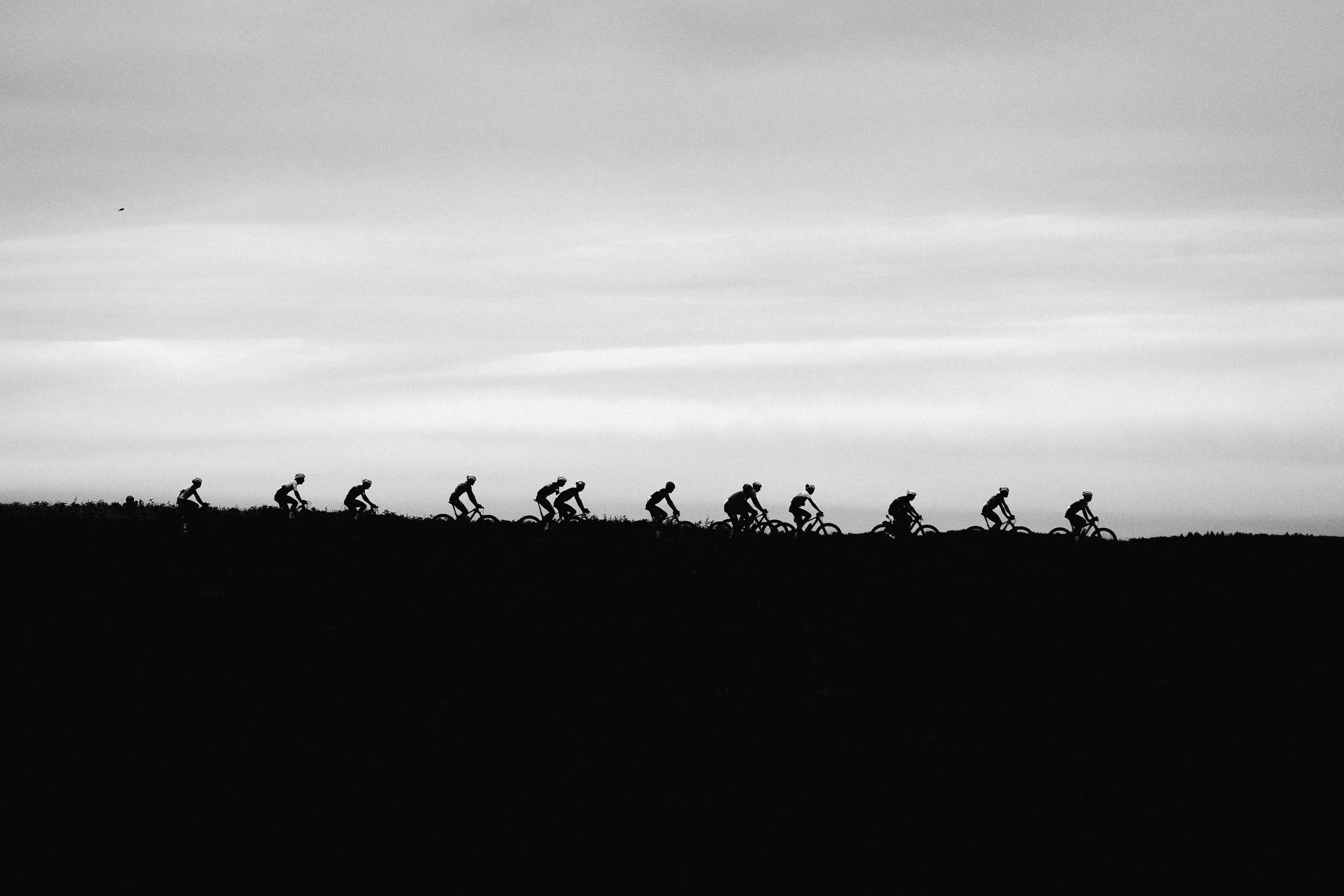 Silhouette of a group of cyclists riding uphill during sunset or sunrise, with a partly cloudy sky in the background.