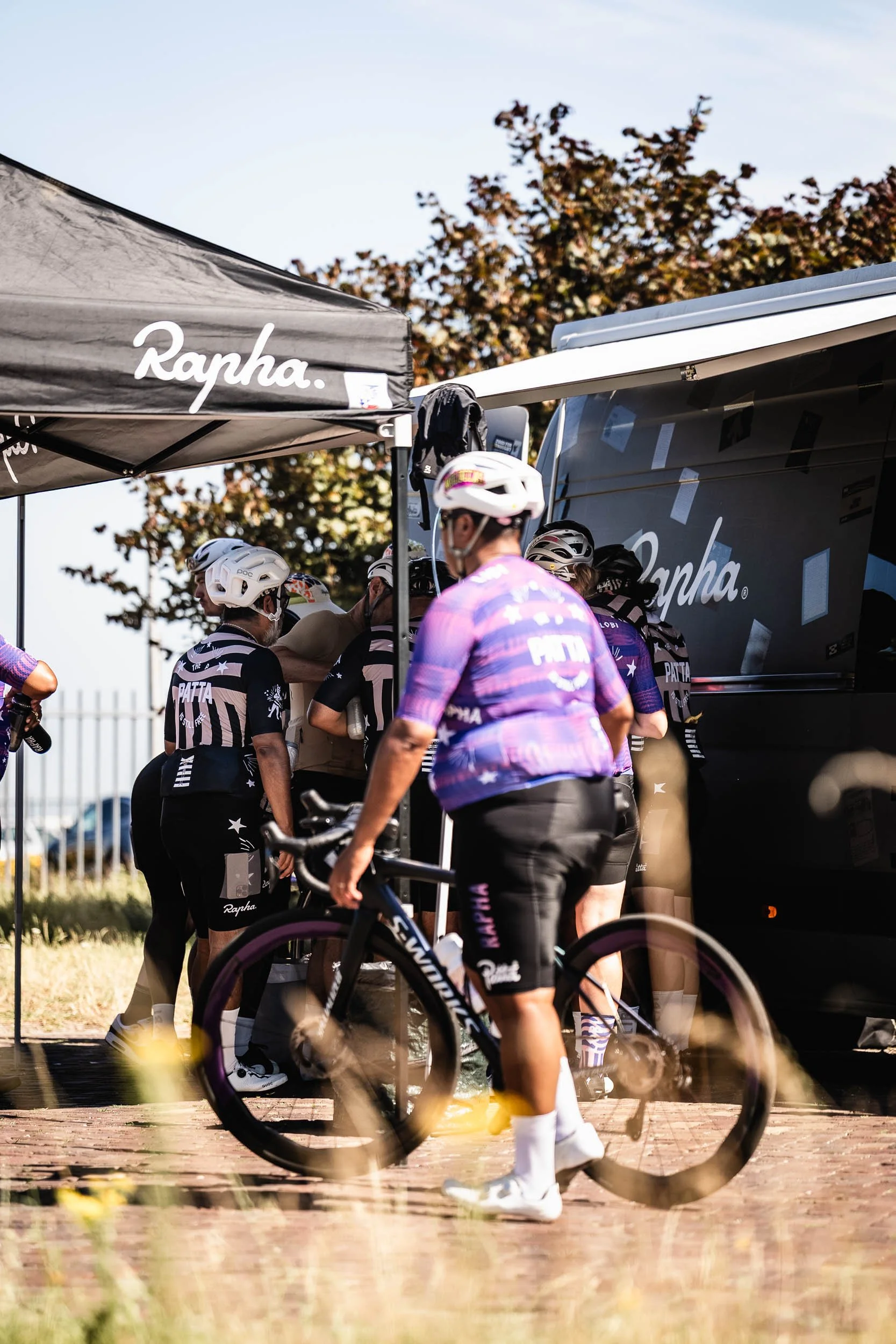 Cyclists in team uniforms and helmets gather near a support vehicle and canopy branded 'Rapha' during a cycling event. (Patta.nl Rapha)