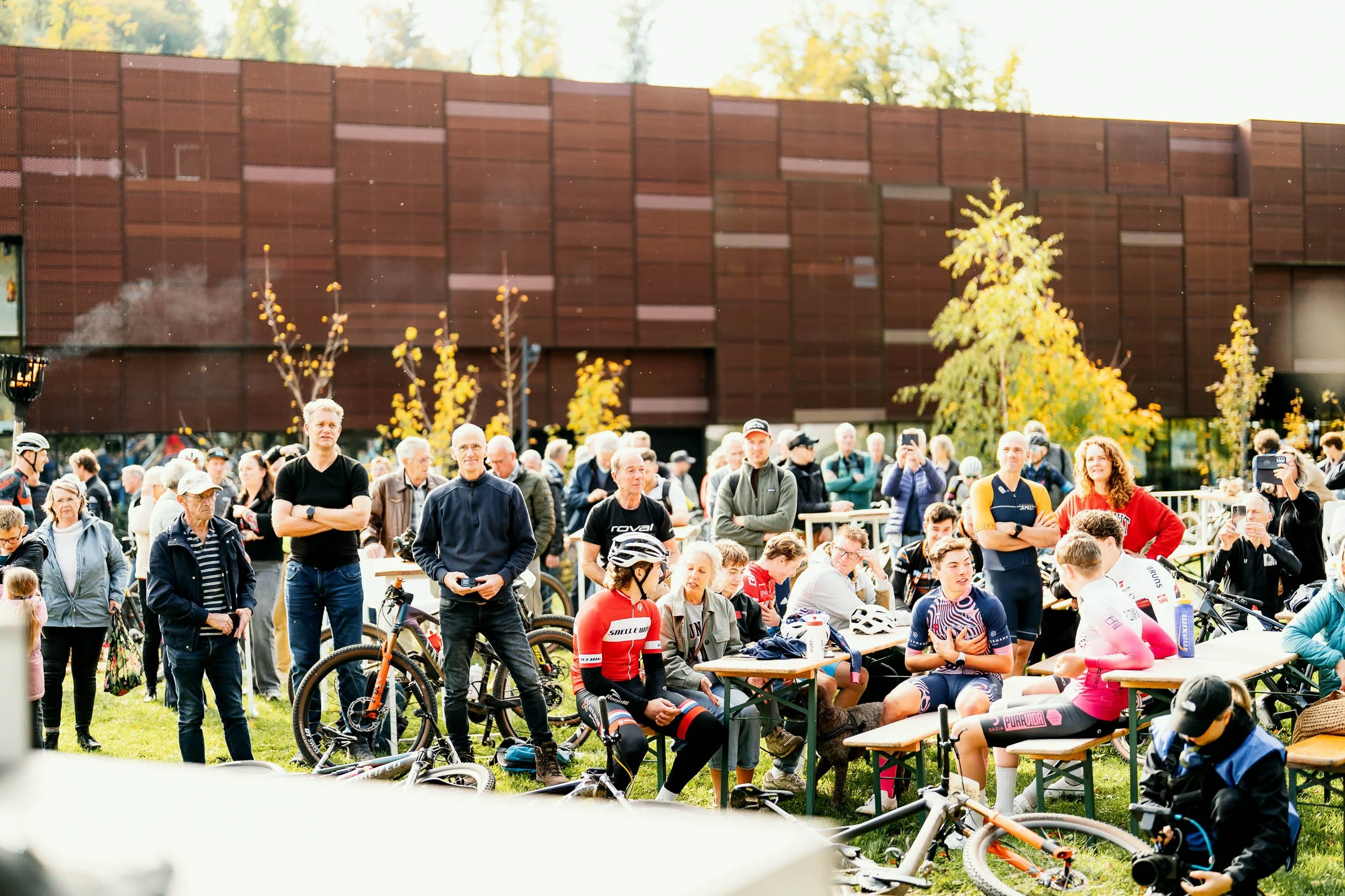 A group of cyclists and spectators gathered outdoors in front of a modern building with brown panels. The scene is lively with some people sitting at tables, others standing, and many wearing cycling gear and helmets.