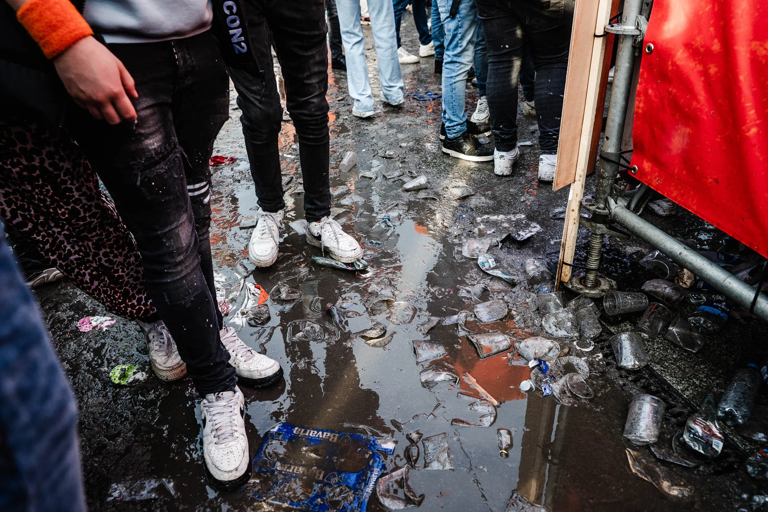 Crowd of people standing on a wet, messy street with broken glass, plastic bottles, and debris scattered around.