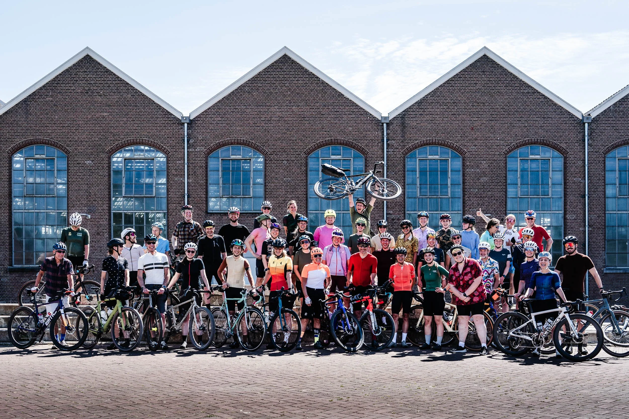 A large group of people standing with bicycles in front of a brick building with large windows. One person is holding a bicycle above their head. Everyone is wearing helmets and dressed in casual or athletic clothing, celebrating together.