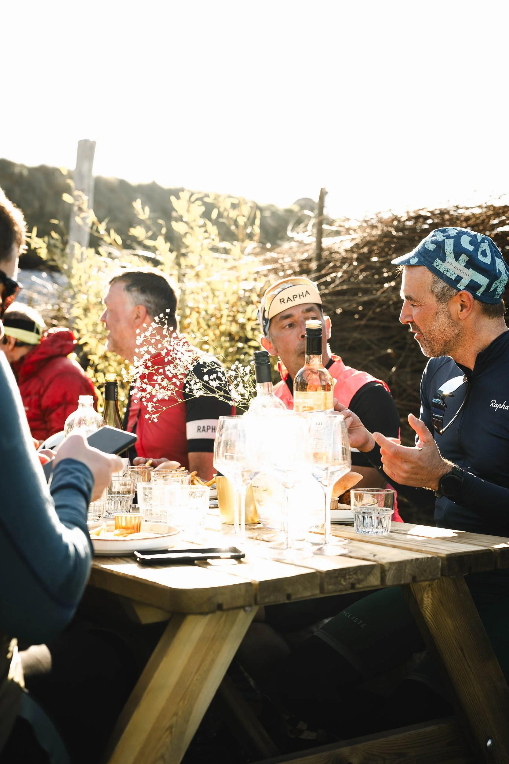 Group of people sitting at a wooden outdoor table, having drinks and conversations, with sunlight and trees in the background.