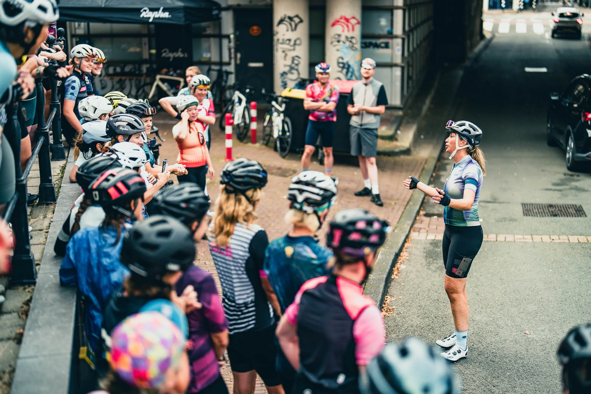 A group of cyclists in helmets gathered around a woman giving instructions outside a building.