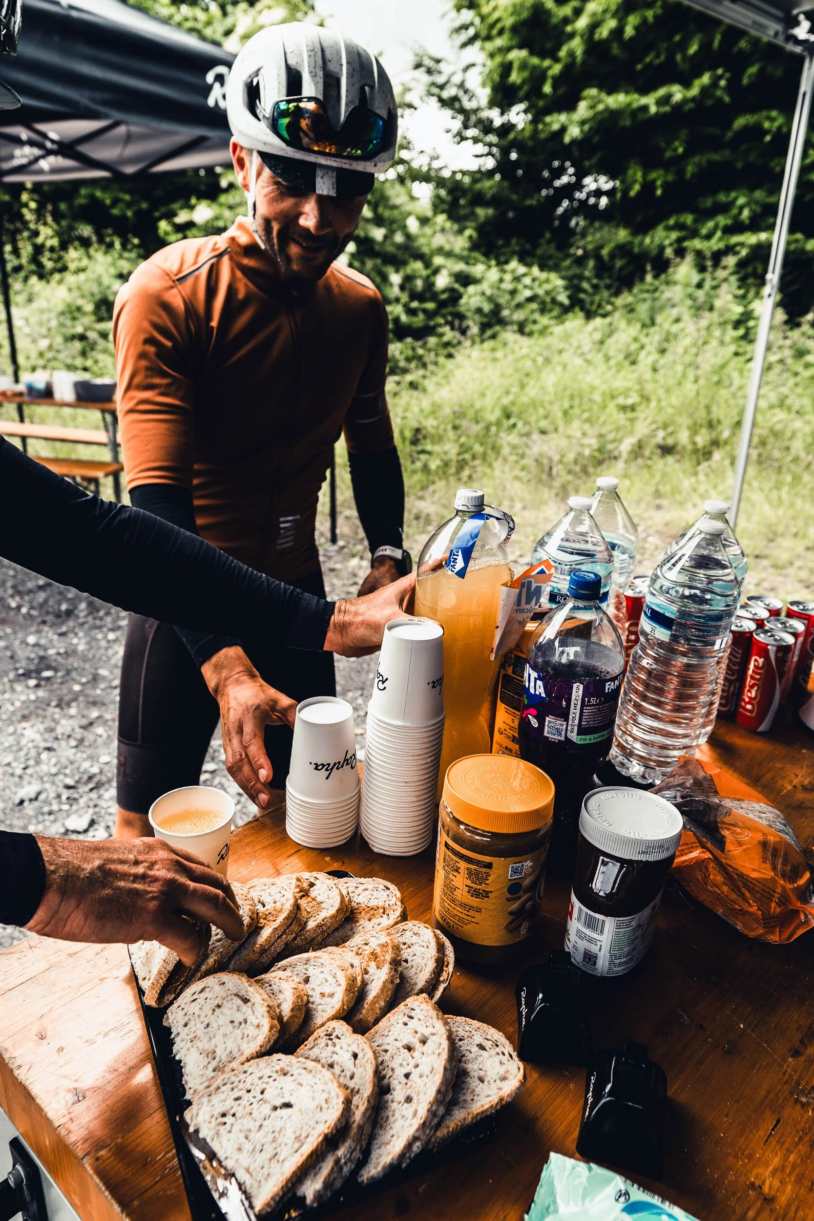 A man in cycling gear and helmet at an outdoor refreshment table with bread, bottled drinks, canned beverages, cups, and snacks, in a wooded area.
