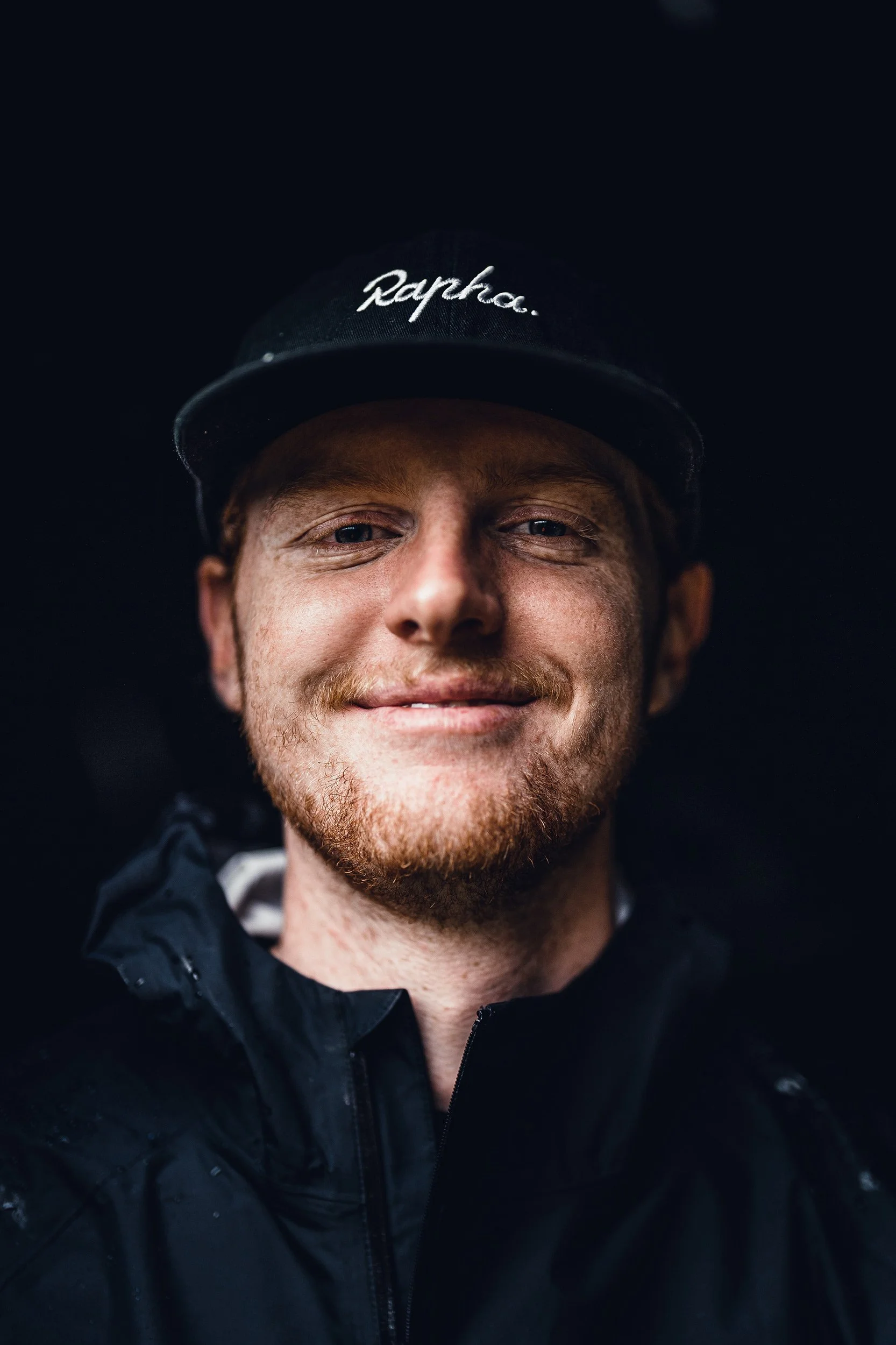 Close-up portrait of a smiling man with red hair and beard wearing a black cap that says 'Rapha' and a black jacket.