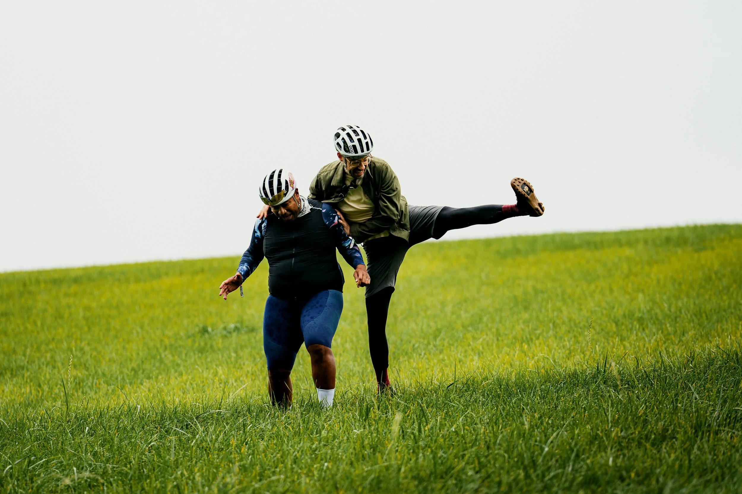 Two men wearing cycling helmets are playfully wrestling on a grassy hill. One man is lifting his leg in the air as the other man leans forward.