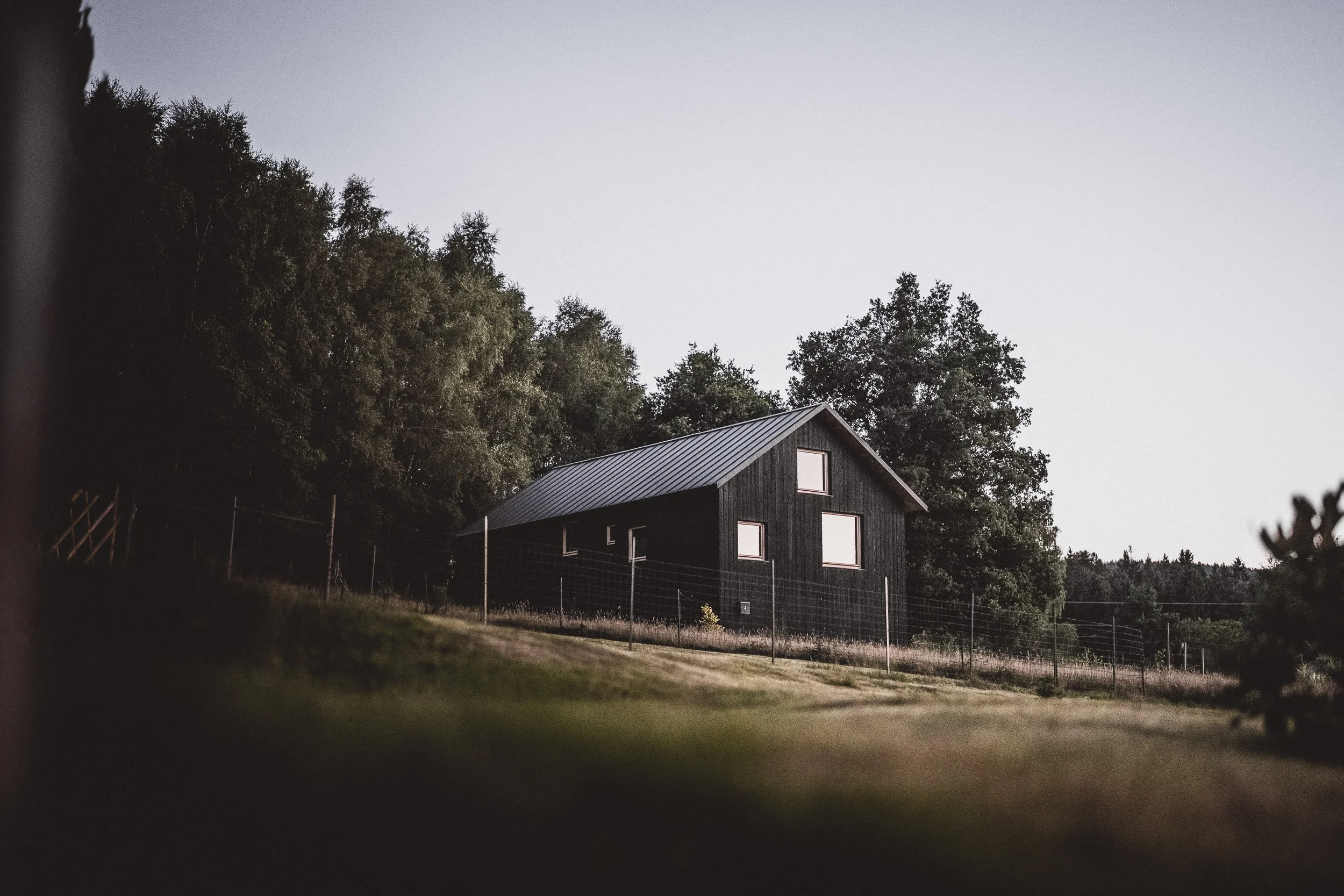 A black wooden house with a metal roof situated on a grassy hill, surrounded by trees, with a wire fence in front, during dusk or dawn.
