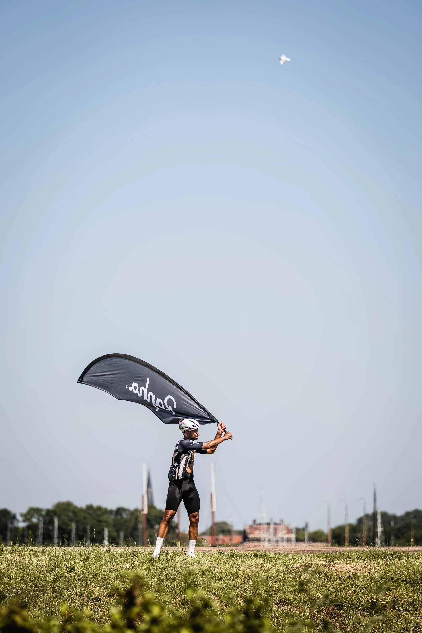 A cyclist in athletic gear, helmet, and sunglasses holding a black flag with white text, standing on a grassy field with clear sky and plane flying overhead in the background. (Patta.nl Rapha)
