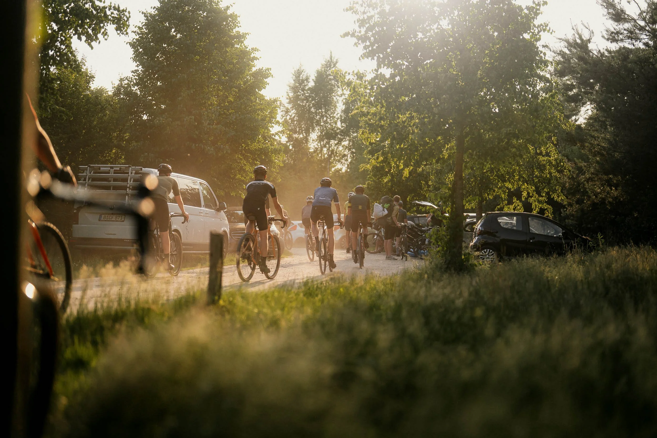 Group of cyclists riding on a dirt path surrounded by parked cars and trees in warm sunlight. (Graveller) )(Canyon)