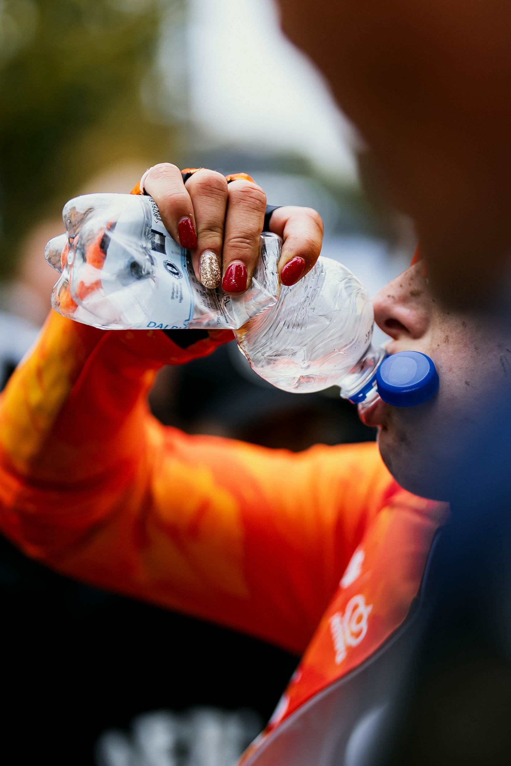 A person drinking water from a plastic bottle during outdoor activity, wearing an orange and black sports shirt, with painted red and glittery nails.