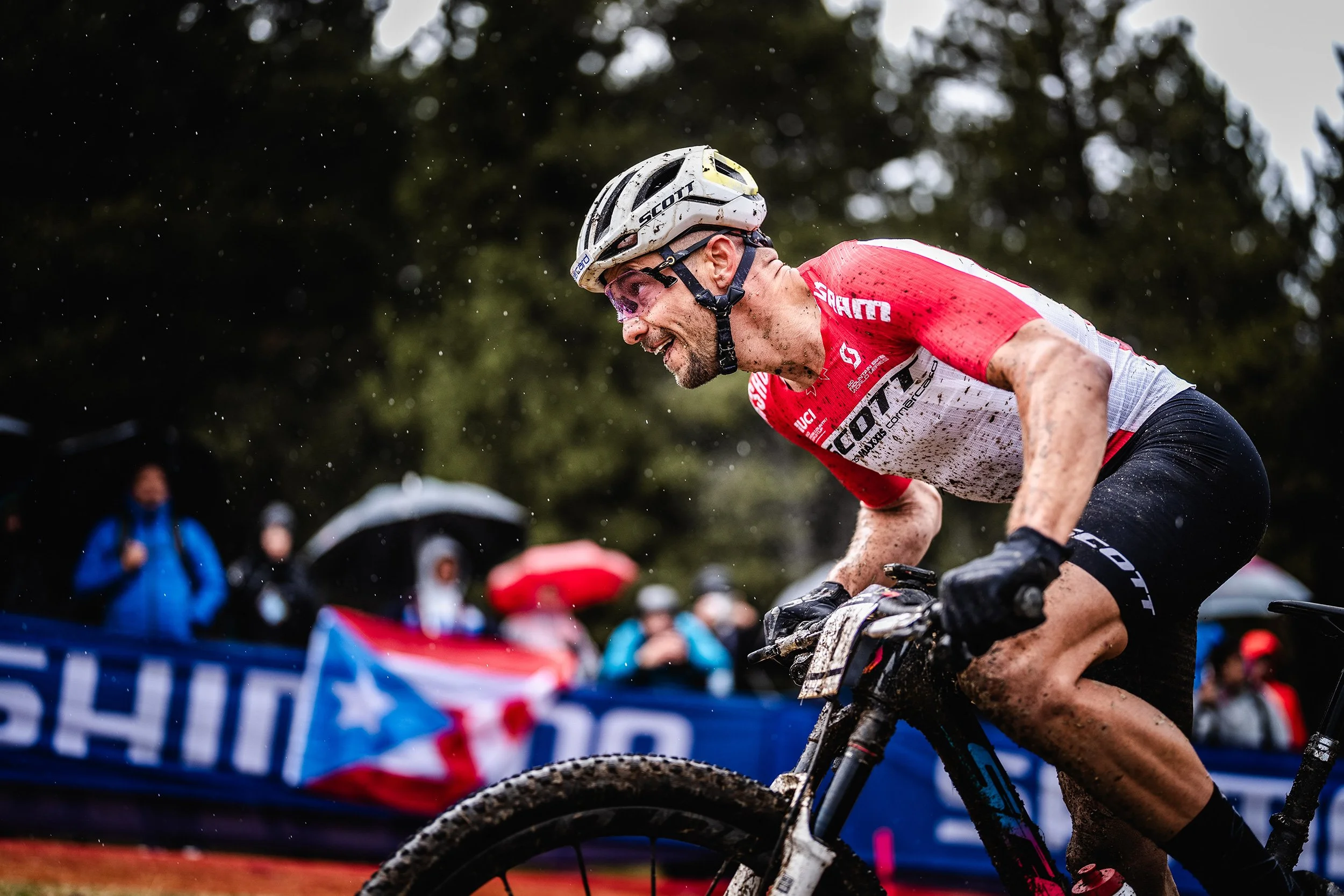 A cyclist in a red and white jersey riding a mountain bike during a race, with mud on his face and body, wearing a helmet, as spectators with umbrellas watch in the background.