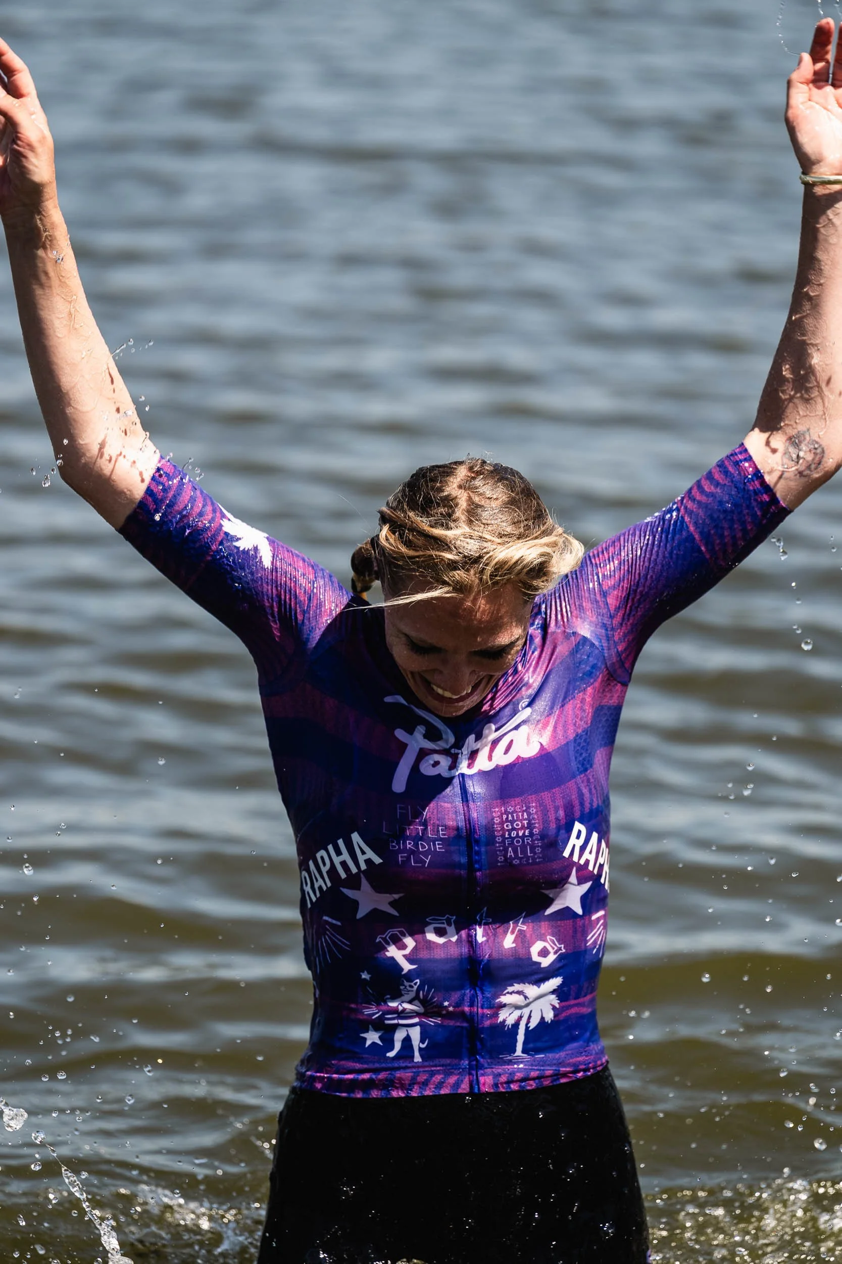 A smiling woman is joyfully splashing water with her arms raised in a body of water, wearing a colorful cycling jersey. (Patta.nl Rapha)
