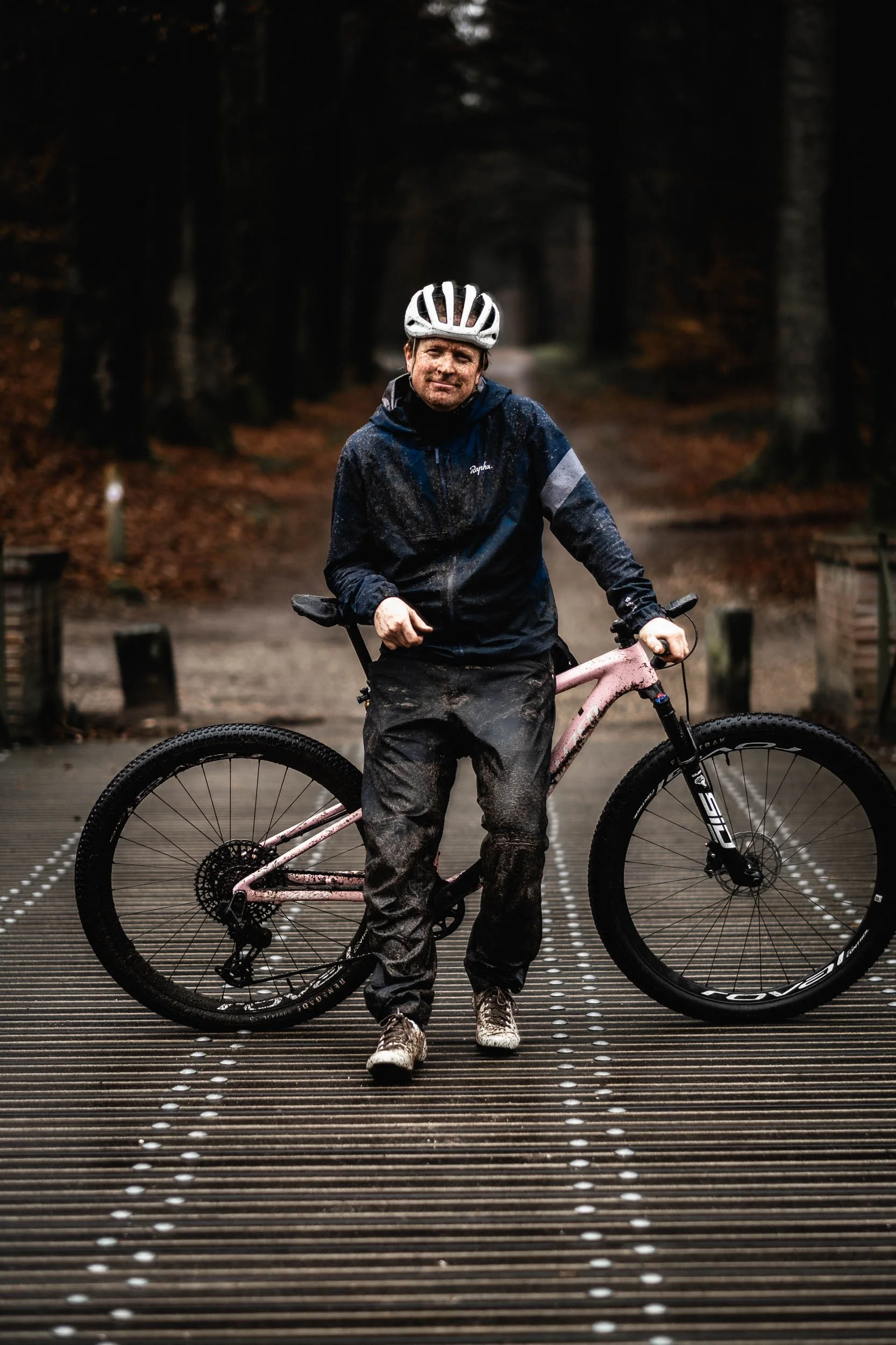 Person standing on a wet bridge with mountain bike in outdoor forest setting, wearing a helmet and rain gear. (Vincent Engel Fotografie)