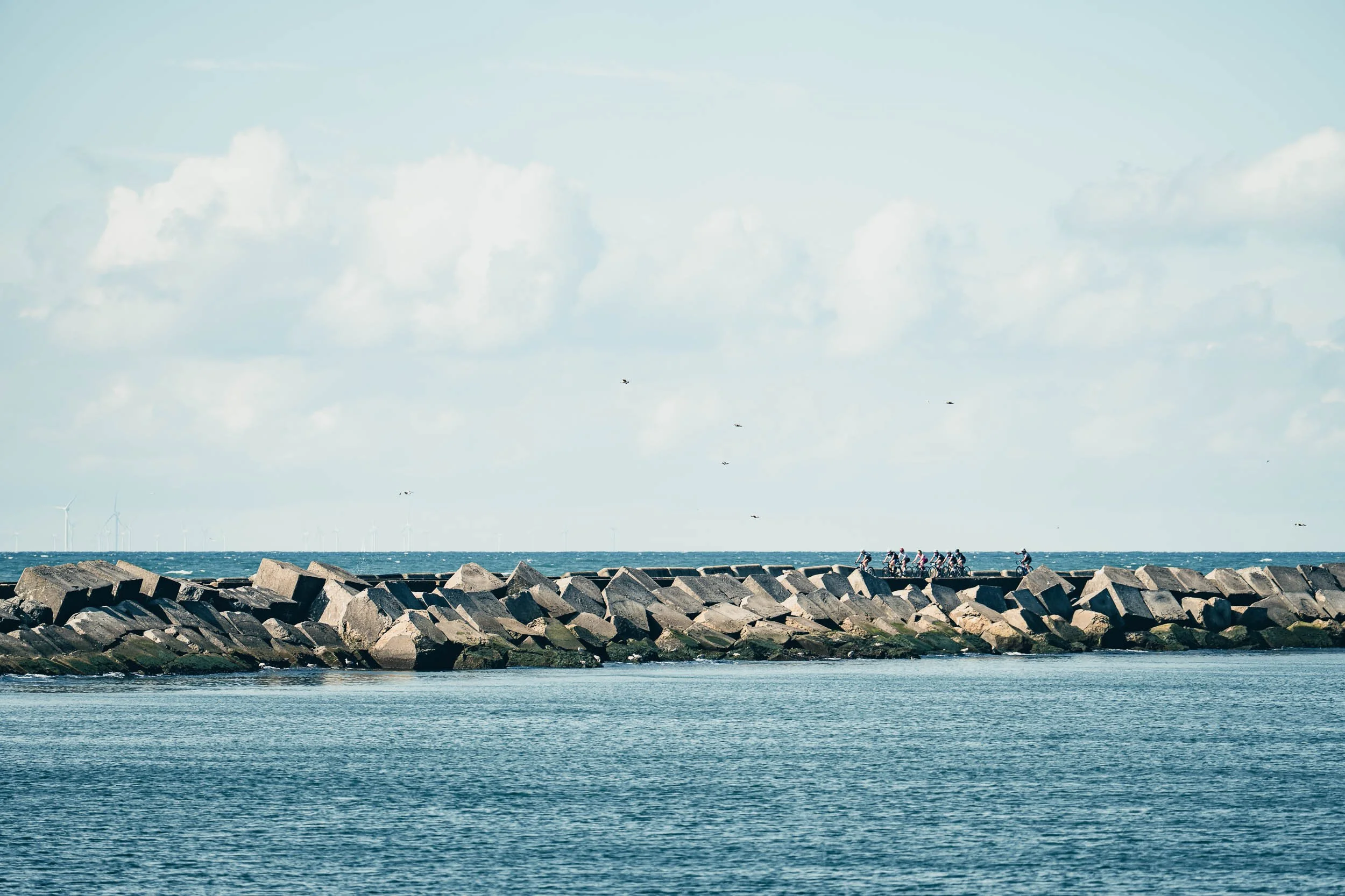 A seascape with large rocks forming a breakwater, a group of cyclists riding on a path, wind turbines on the horizon, and a partly cloudy sky.