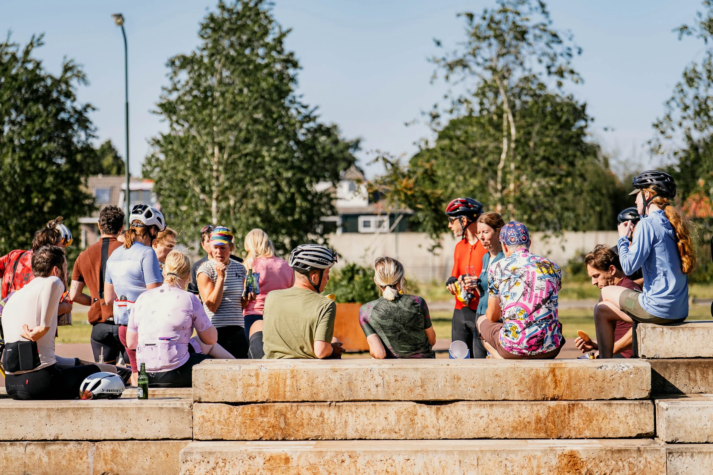 A group of people, some wearing bicycle helmets, gathered outdoors on stone benches and steps, enjoying a sunny day. Some are sitting, some are standing, holding drinks, and chatting, with trees and houses in the background.