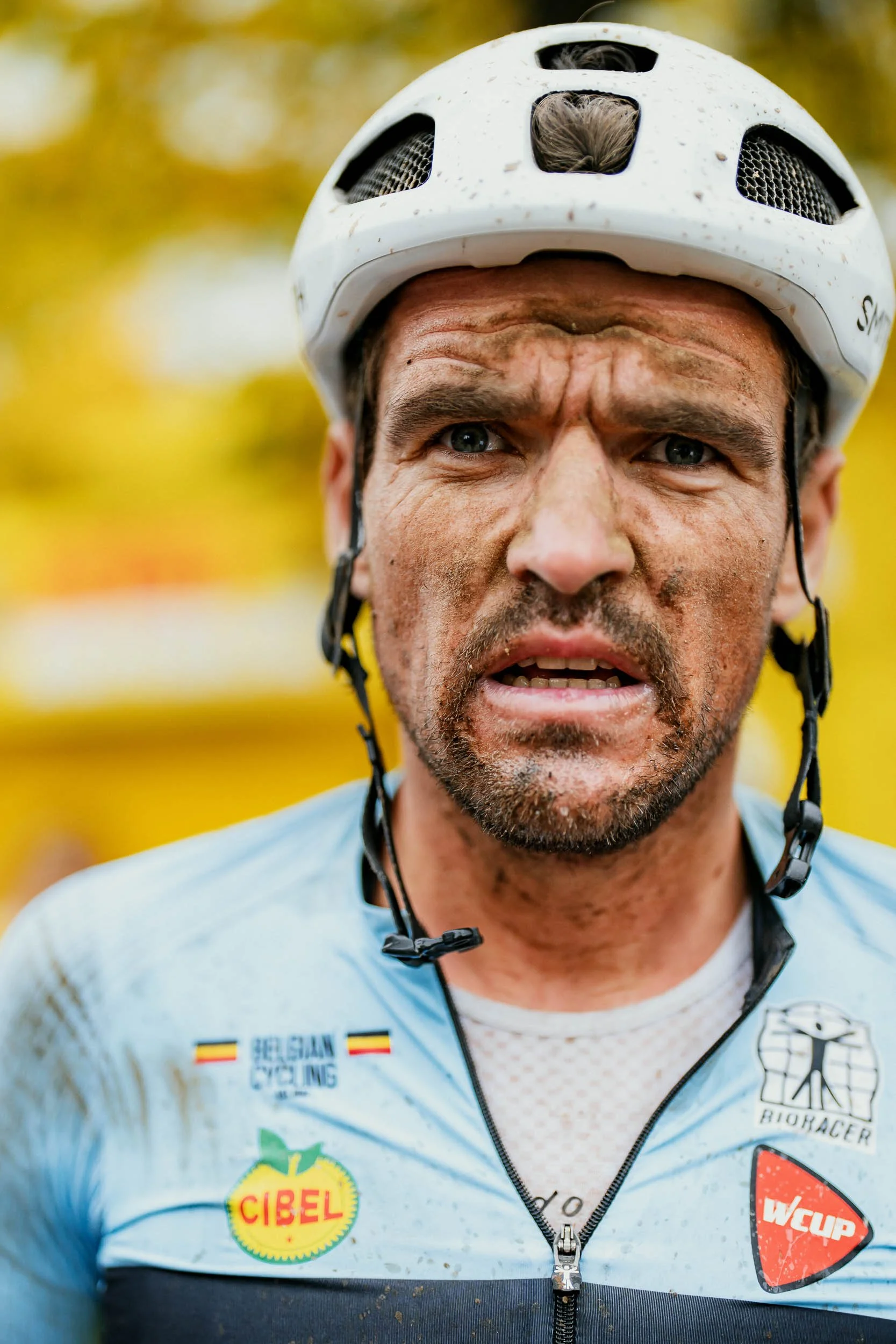 Close-up of a male cyclist with mud on his face, wearing a white helmet and a light blue cycling jersey with various logos.