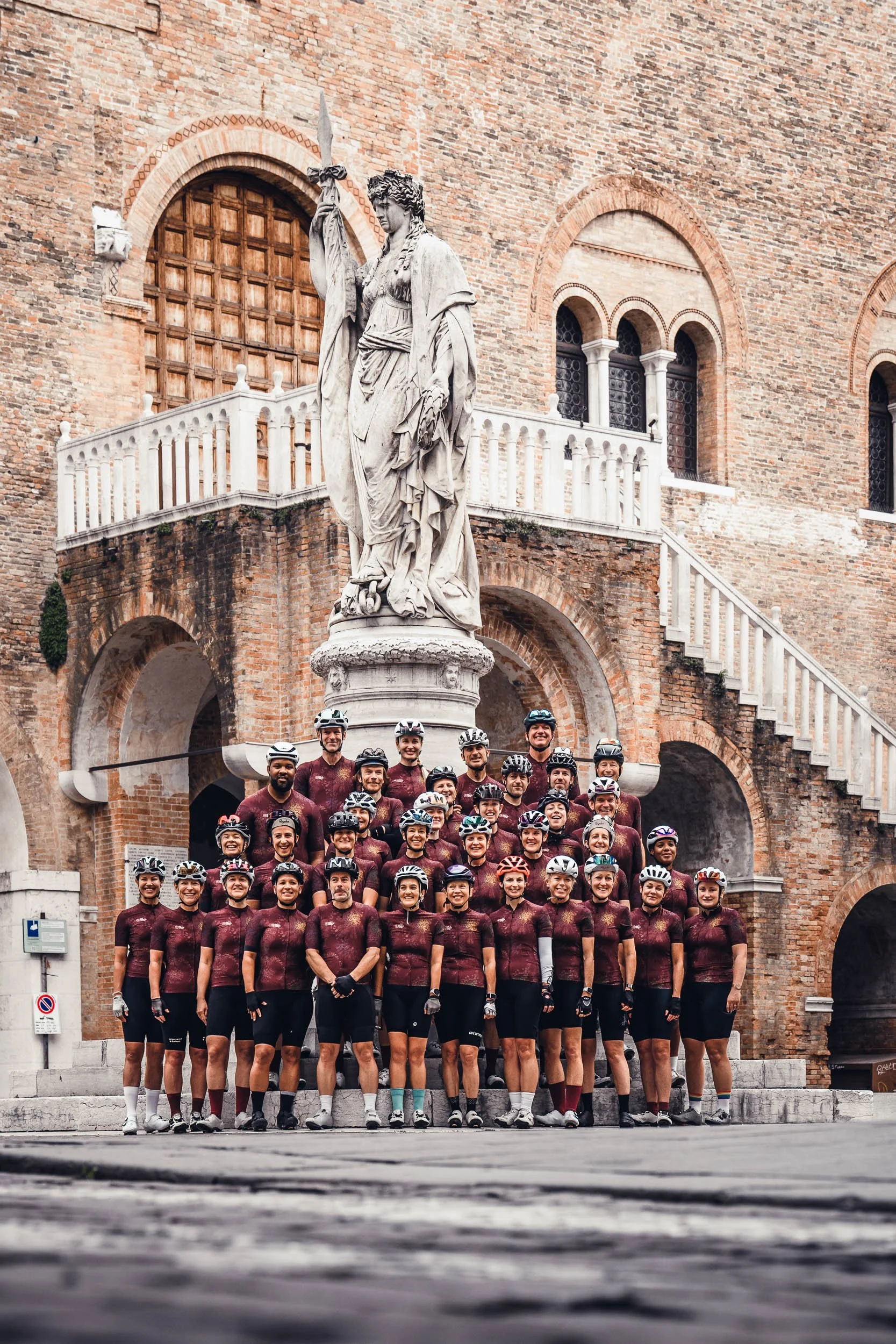A group of cyclists in matching jerseys and helmets standing in front of a large statue of a woman holding a spear, with a brick building in the background.