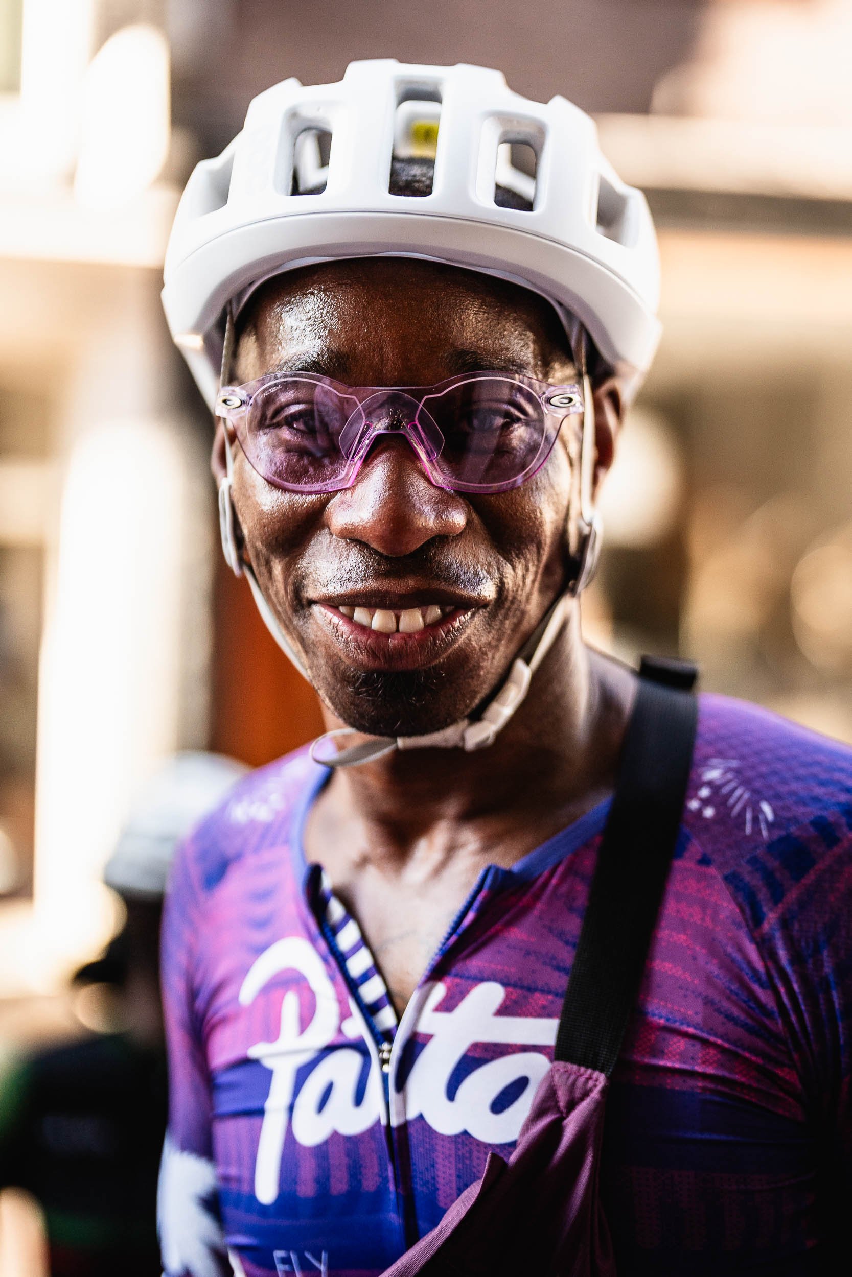A smiling woman wearing a white bicycle helmet, purple-tinted sunglasses, and a purple cycling jersey with white and pink accents. (Patta.nl Rapha)