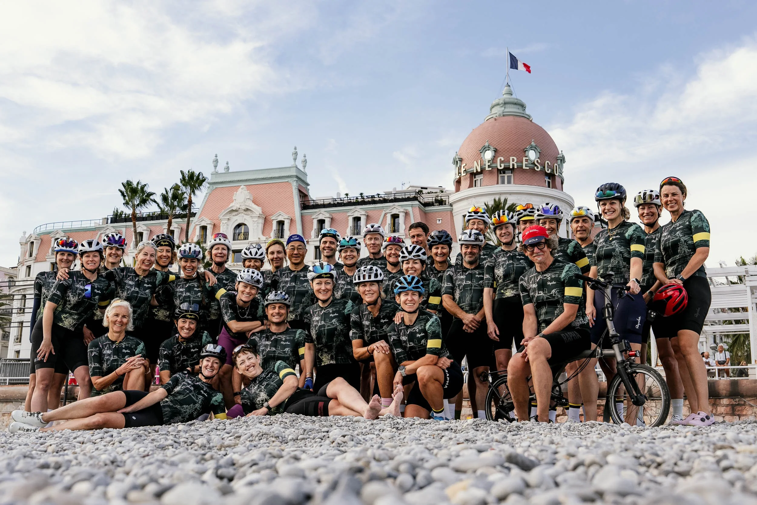 Group of cyclists in black jerseys posing in front of a pink and white building with a flag on top and the words "LE NEGRESCO" on the dome. (One More City  Rapha.cc)