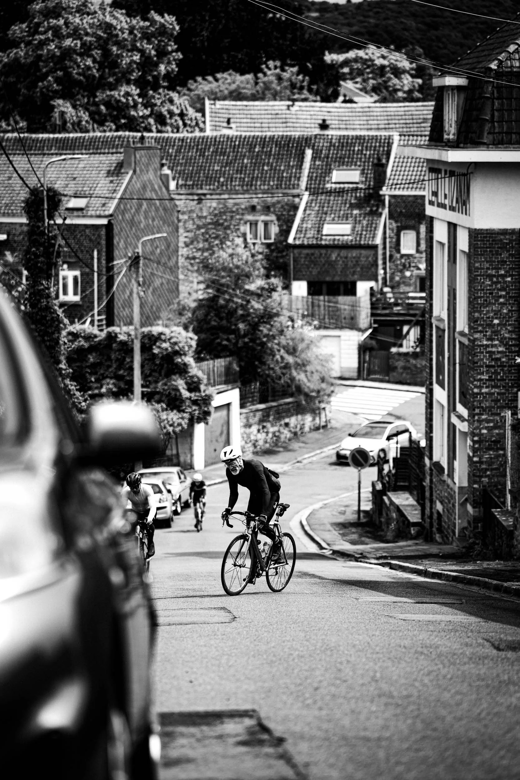 Black and white photo of a downward sloping street with several people riding bikes, wearing helmets, surrounded by residential buildings and parked cars.