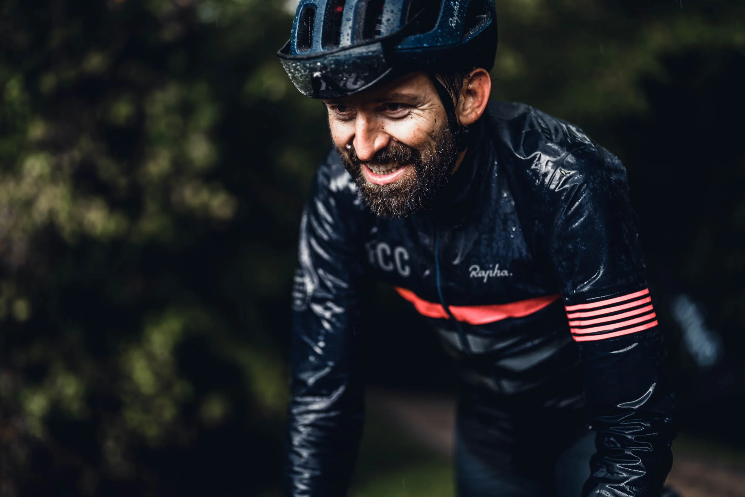 A man in a wet black cycling jersey and helmet, with a beard, smiling while riding his bike outdoors.