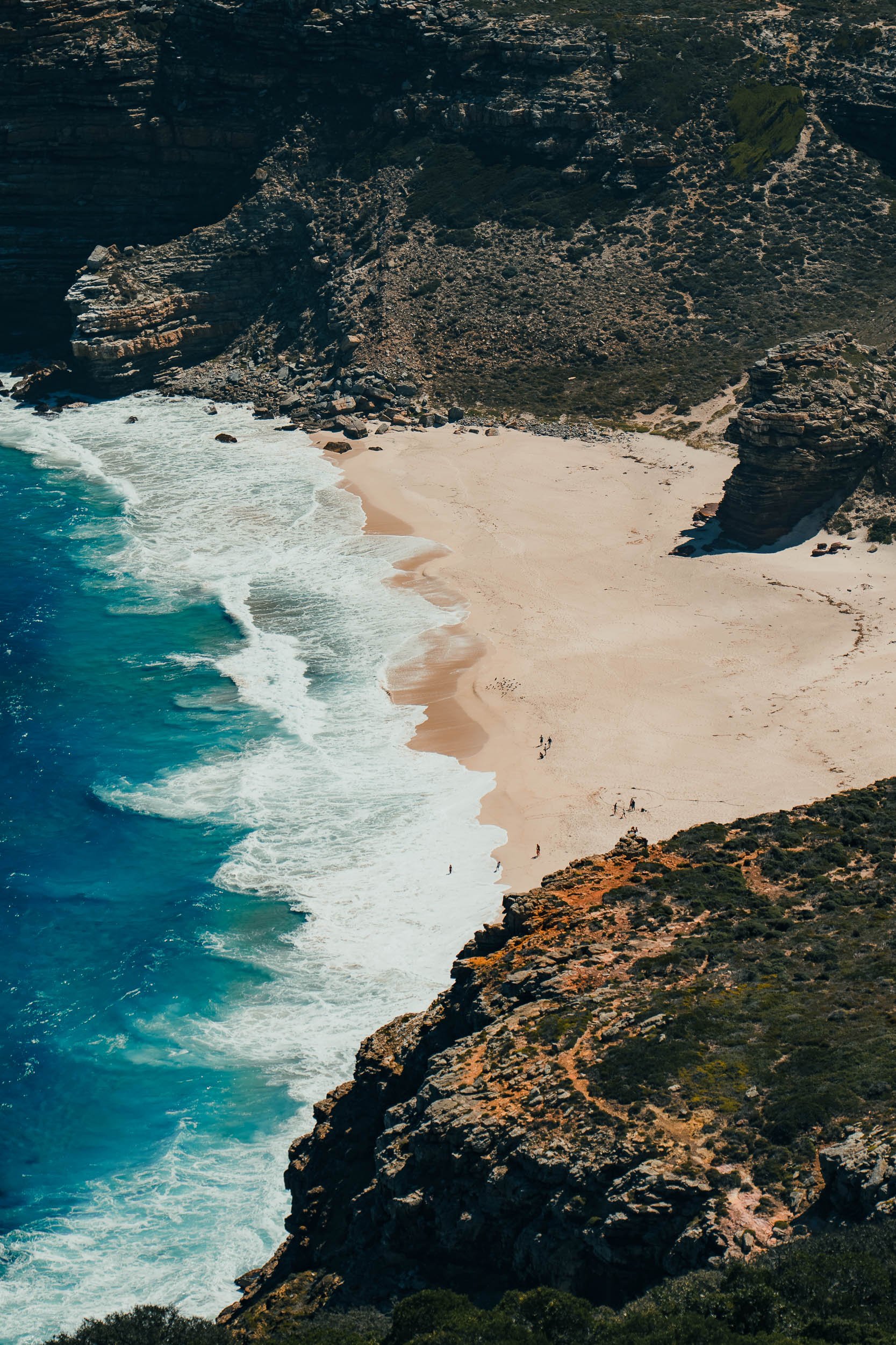 Aerial view of a beach with turquoise ocean waves crashing onto a sandy shoreline, surrounded by rocky cliffs and sparse vegetation.