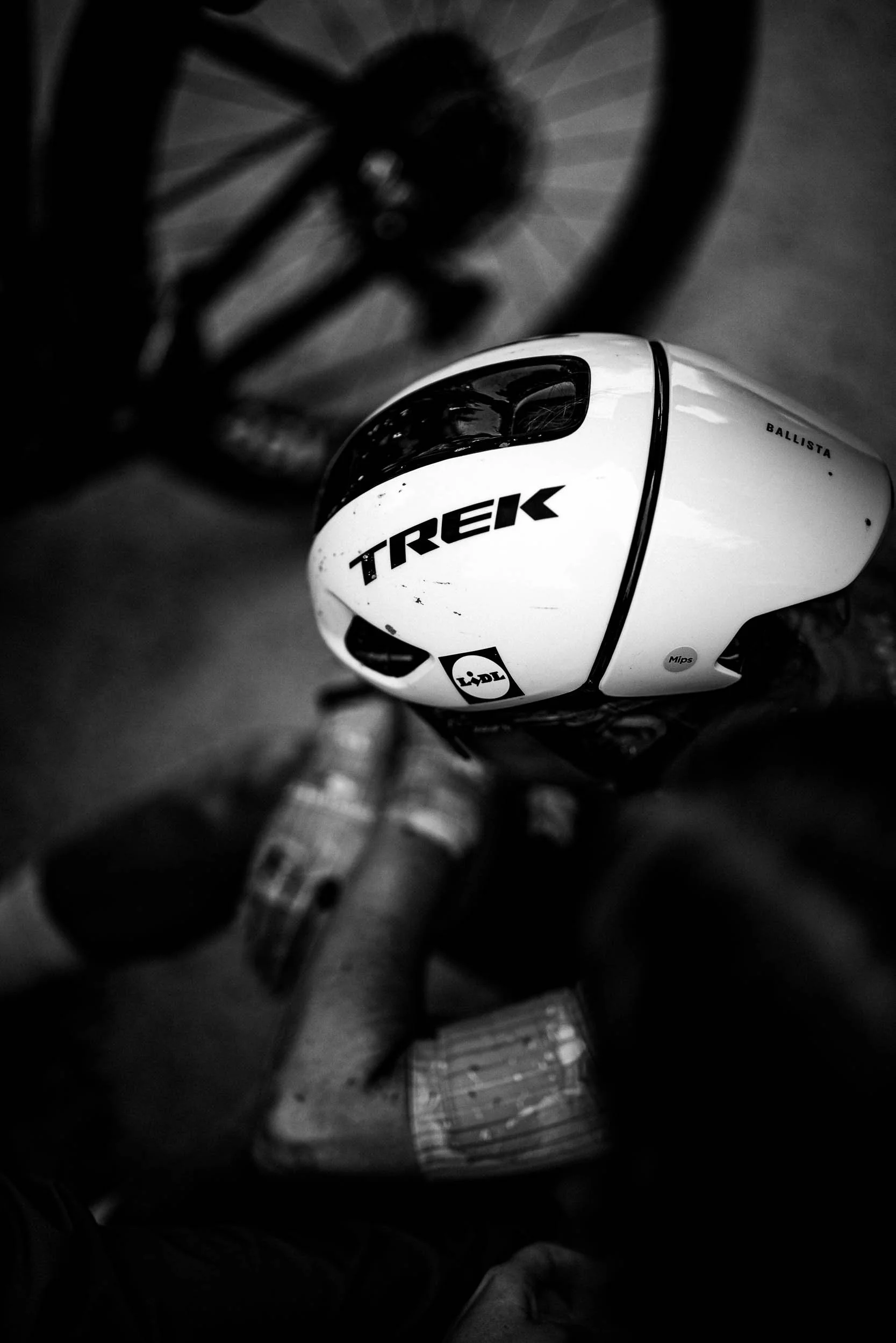 A black and white photo of a cyclist's helmet and handlebar, with a bicycle in the background. The helmet has the brand name 'Trek' on it.