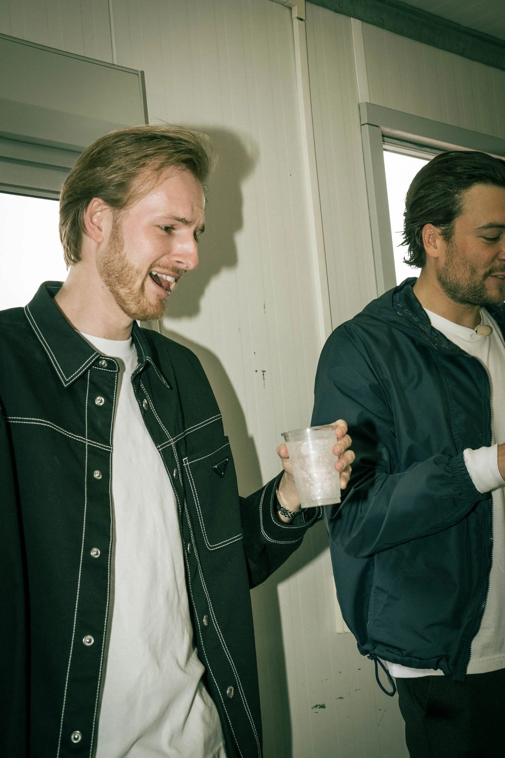 Two men standing indoors, one holding a clear plastic cup with ice, appear to be at a social gathering.   (Viktor Verhulst)