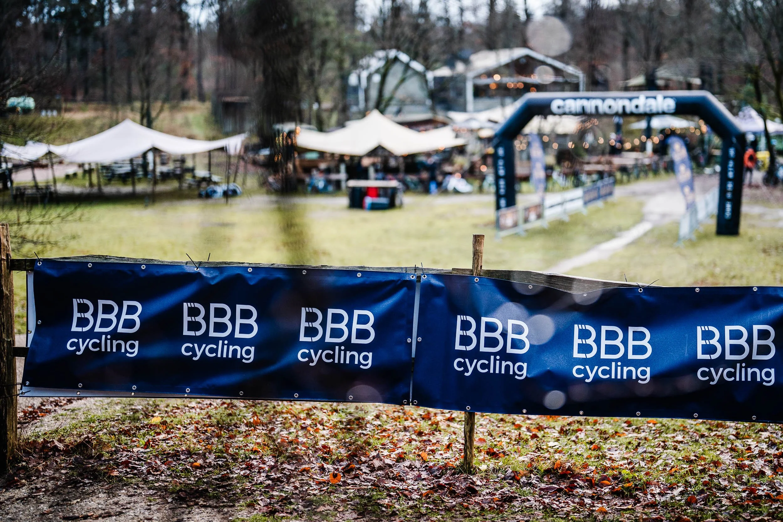 Cycling event with a BBB Cycling banner in the foreground and a muddy pathway leading to a tented area with booths and an air gate in the background on a rainy day.