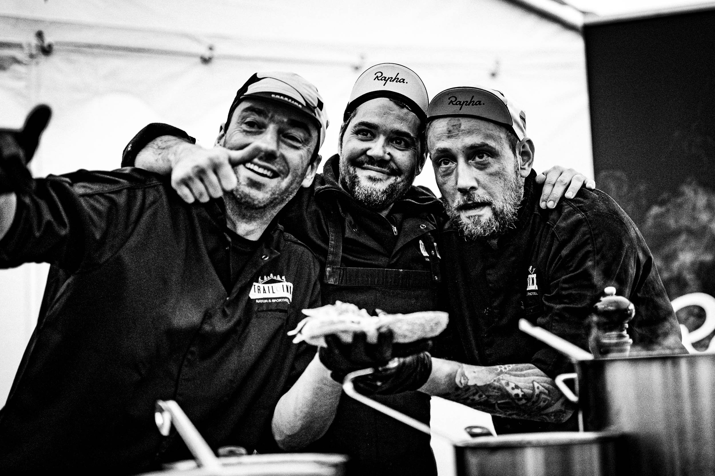 Three men in black jackets and hats, smiling and posing for a photo in a kitchen, with one holding bread, near pots on a stove.