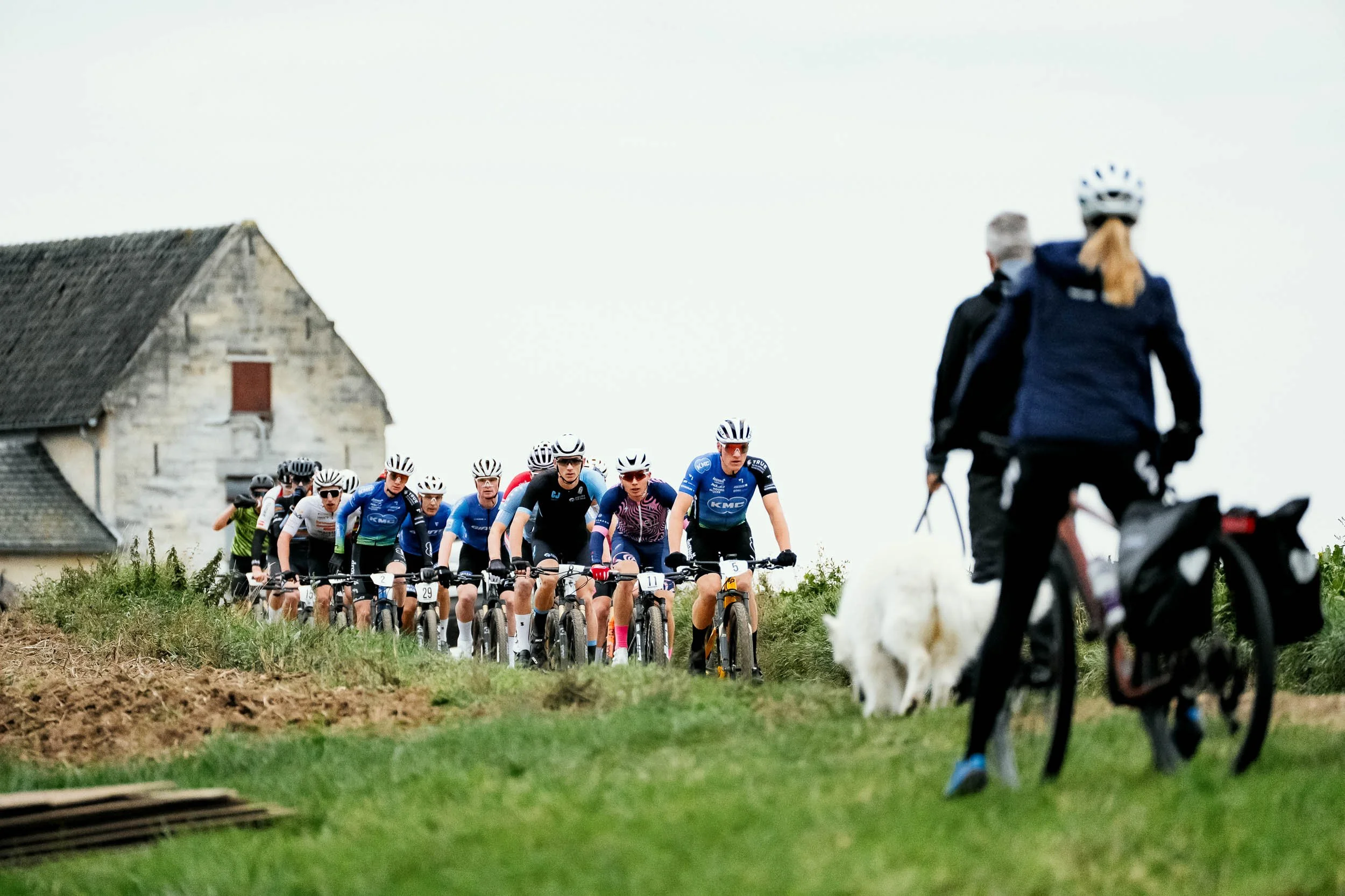 A group of cyclists riding on a rural path with two people on a bicycle and a dog in the foreground, and an old house in the background.