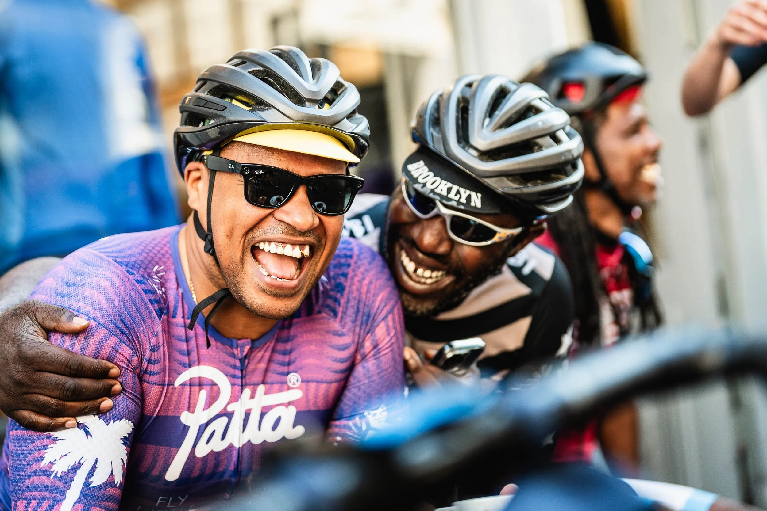 Two men wearing cycling helmets and sunglasses laughing and smiling, with other cyclists in the background. (Patta.nl Rapha)