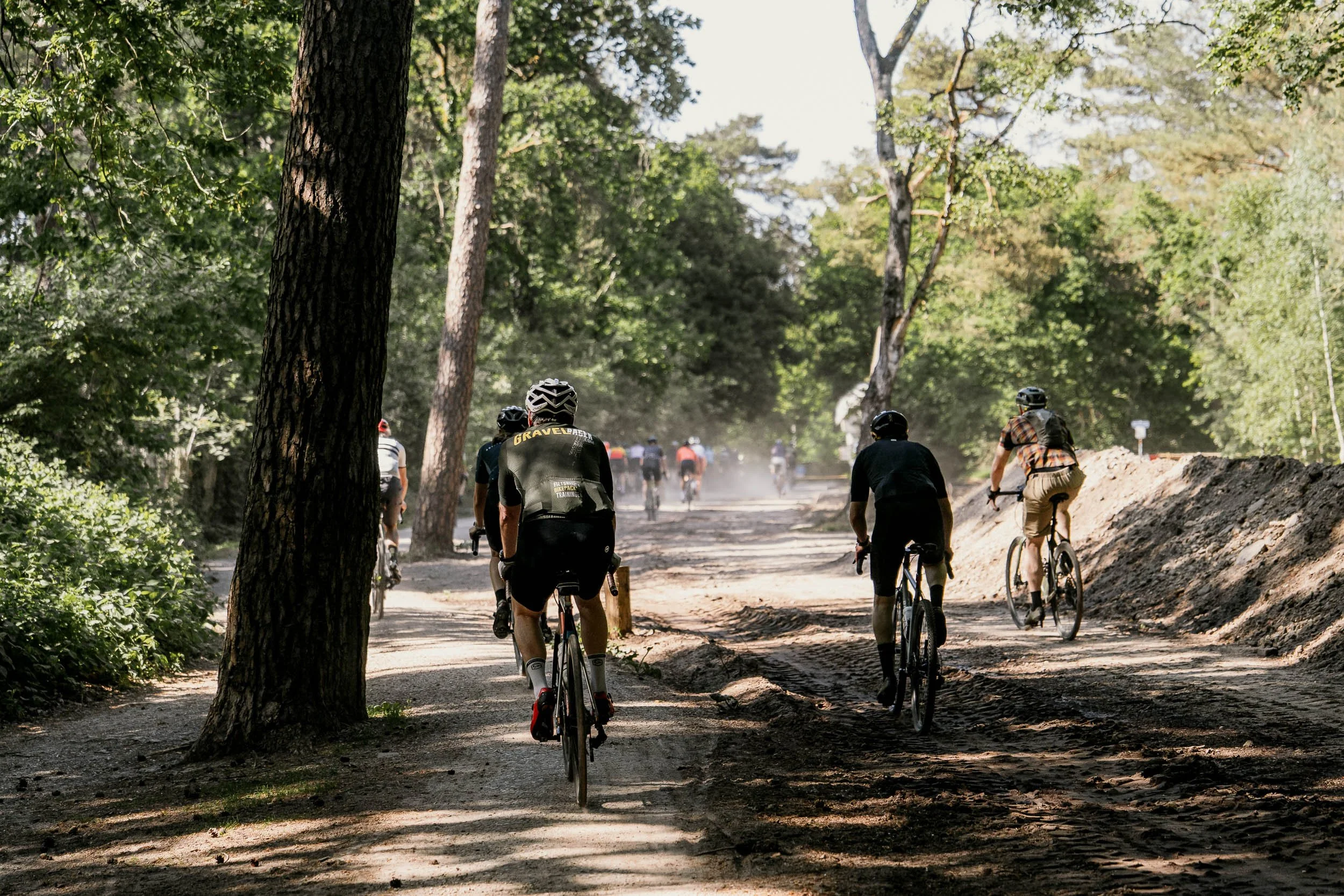 Group of cyclists riding on a dirt trail through a forested area with tall trees and sunlight filtering through the leaves.