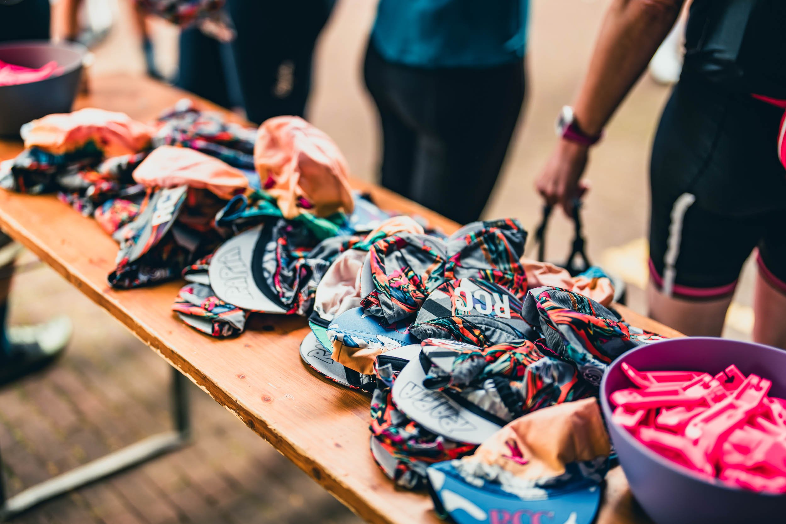 A table with colorful, patterned fabric headbands and visors, some with logos, and a purple bowl filled with pink clips or accessories, likely at a sporting or outdoor event.